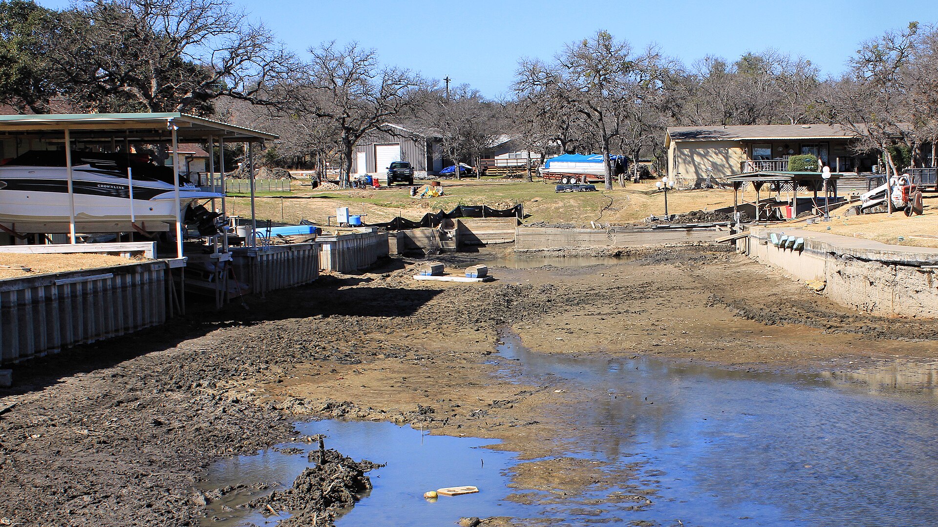 Drained shoreline of Lake LBJ with exposed mudflats, boat docks, and lakeside homes