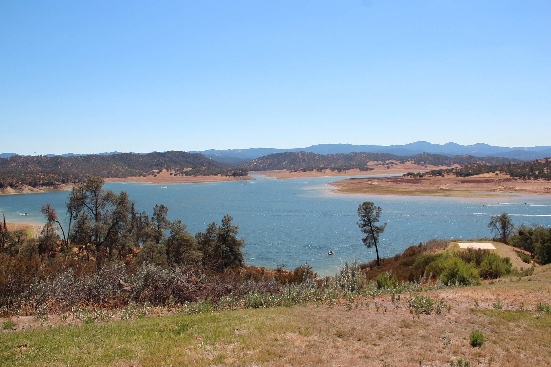 Lake Nacimiento's blue water with oak-covered hills and a grassy shoreline in the foreground.