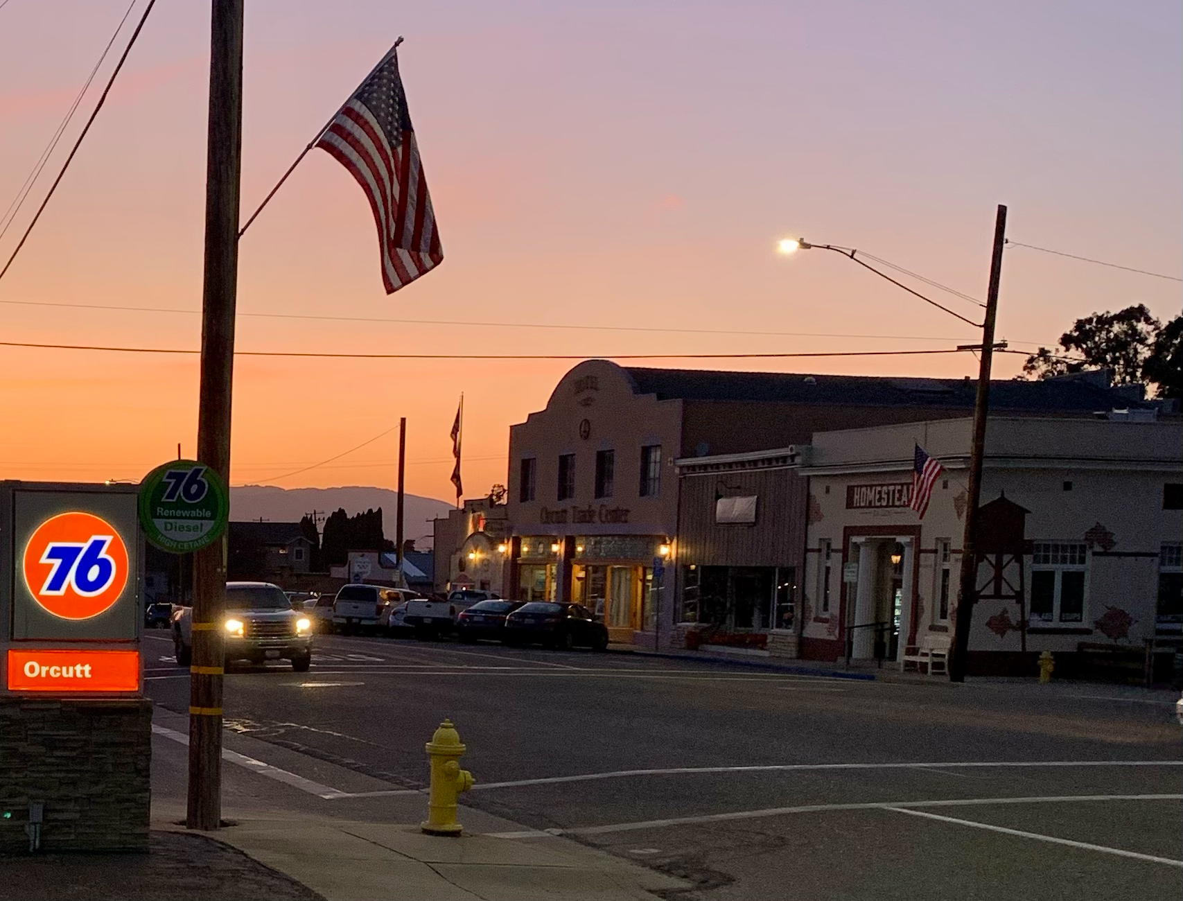 Clark Avenue in Old Town Orcutt at sunset, with storefronts, a gas station sign, and an American flag.