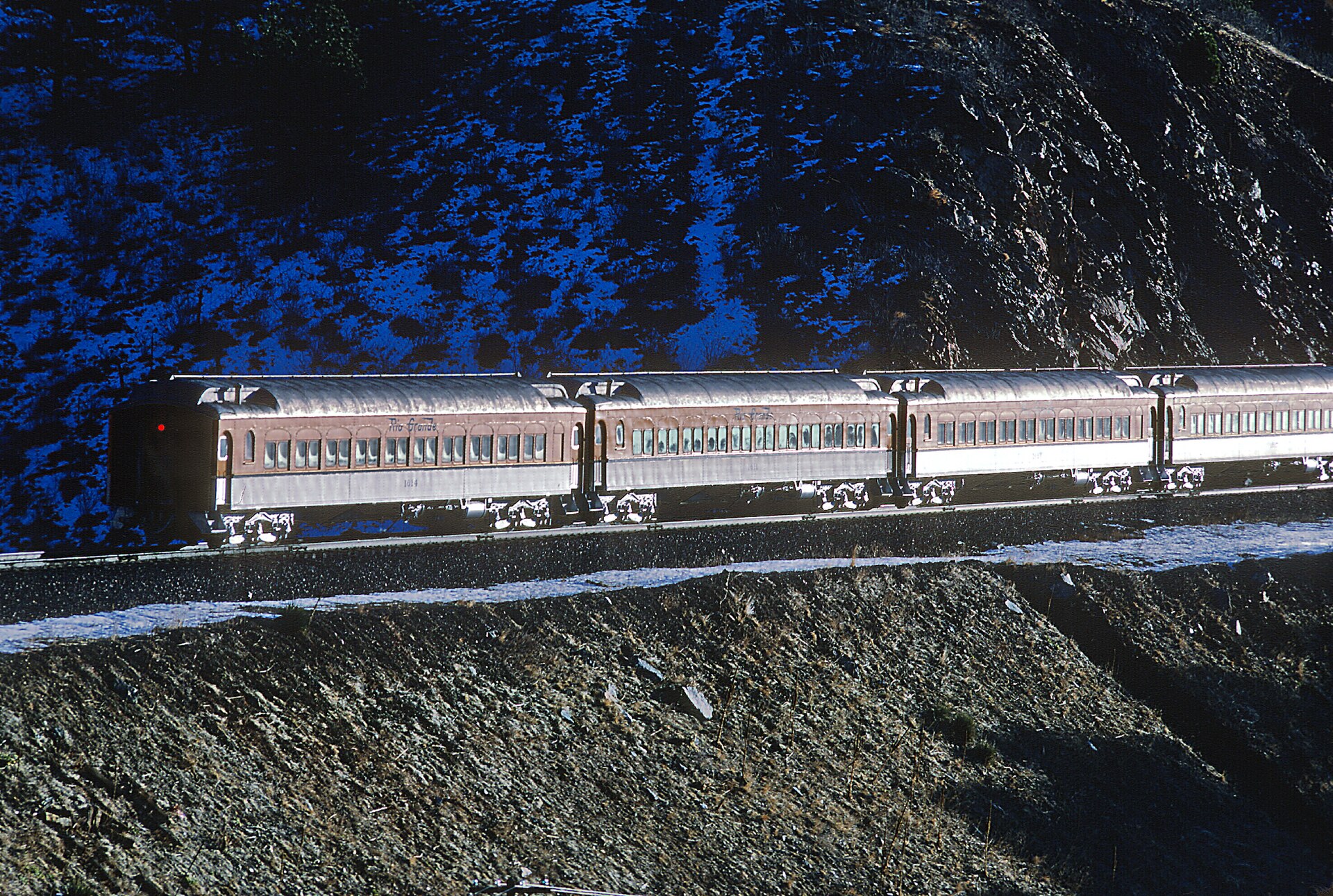 A silver passenger train on snowy tracks in a rocky canyon.