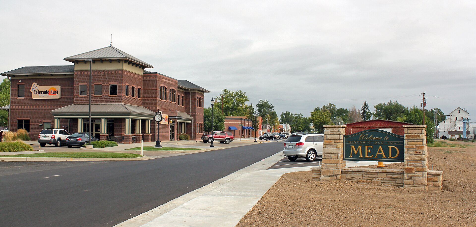 A wide main street in Mead, Colorado, with commercial buildings and a town sign.