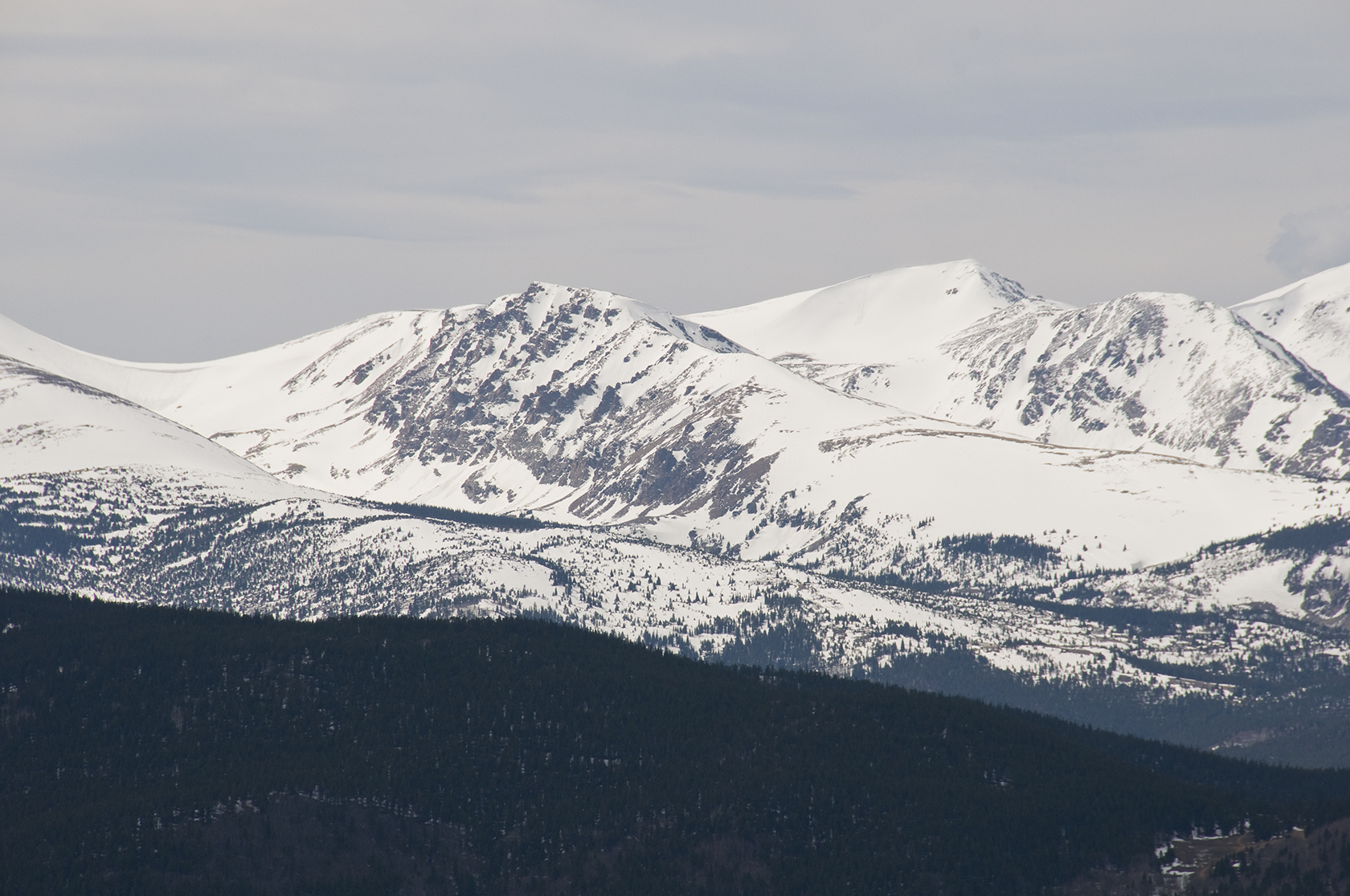 Snow-covered mountain peaks rising above dark forested slopes under a pale overcast sky.