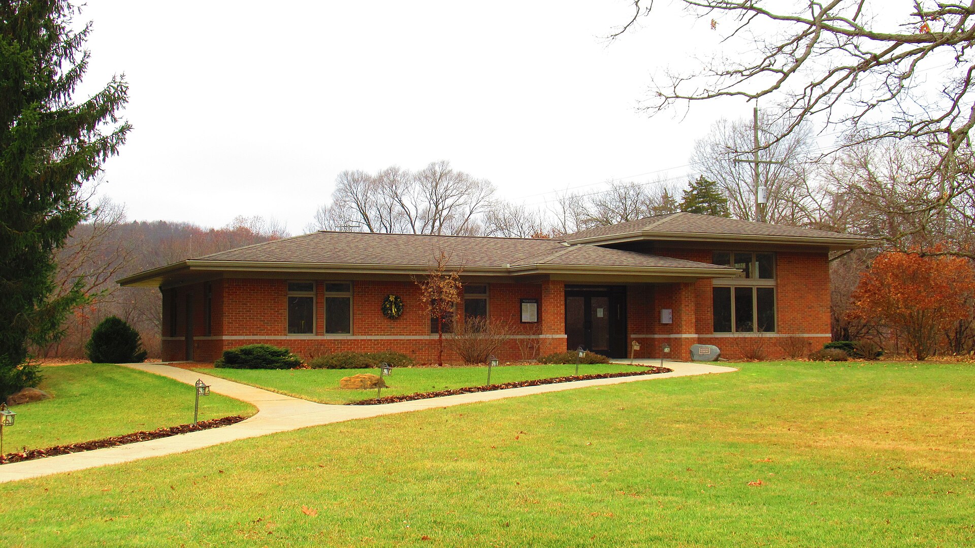 A low brick village hall building with a sloped roof, set on a green lawn with mature trees.