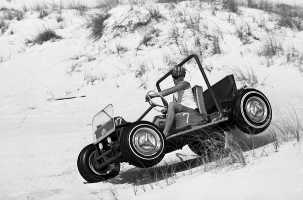 Black-and-white photo of a dune buggy driving on a sandy beach.
