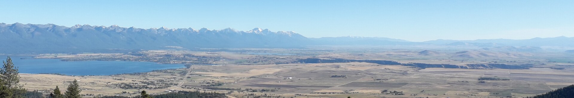 A wide panoramic view of a river valley and lake with jagged mountain peaks on the distant horizon.
