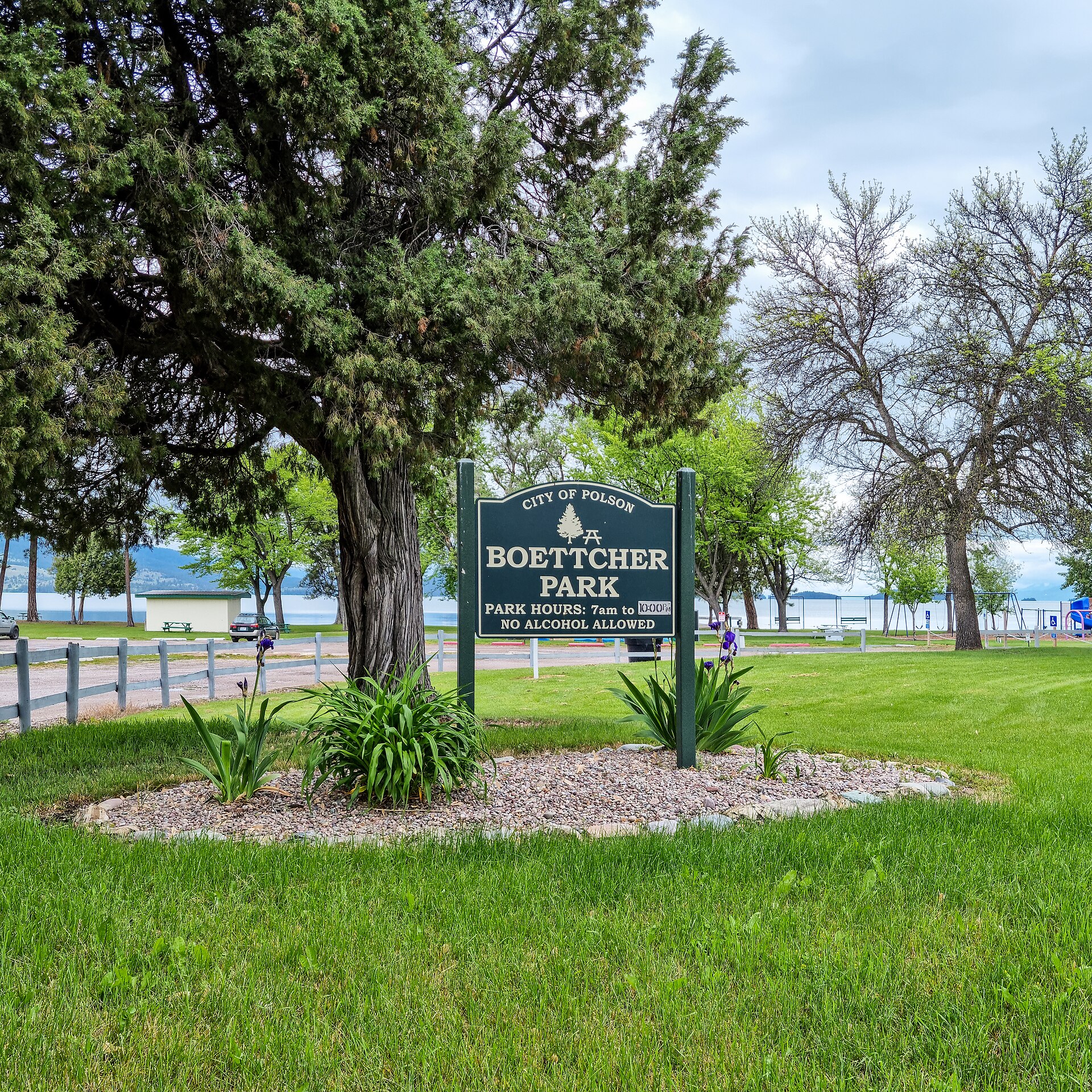 A green park sign reading Boettcher Park on a lawn with trees and a fence behind it.