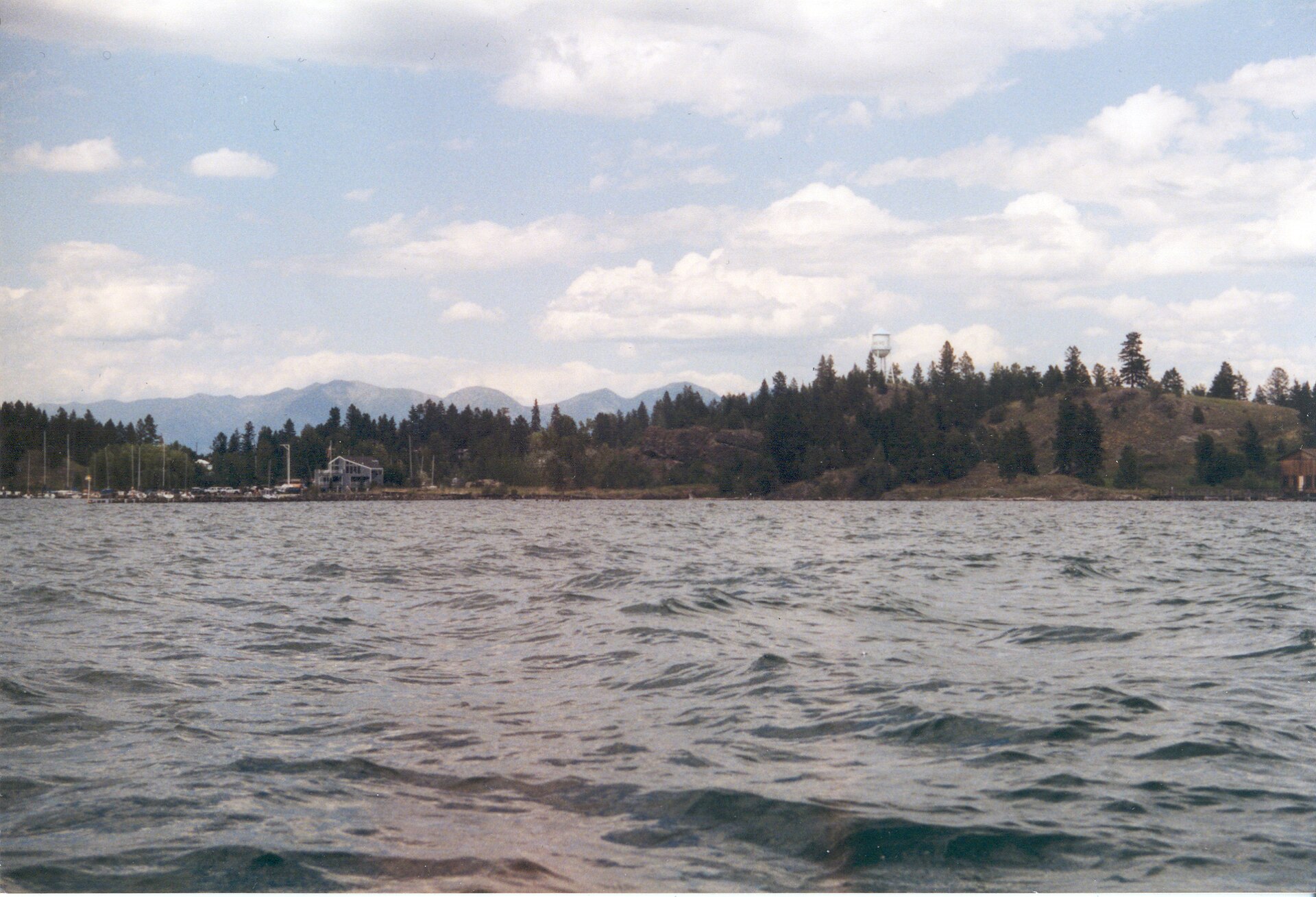Choppy lake water in the foreground with a tree-lined shore, marina buildings, and mountains behind.