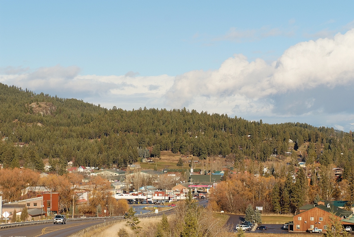 A small-town street with low buildings set against forested hills under a bright blue sky.