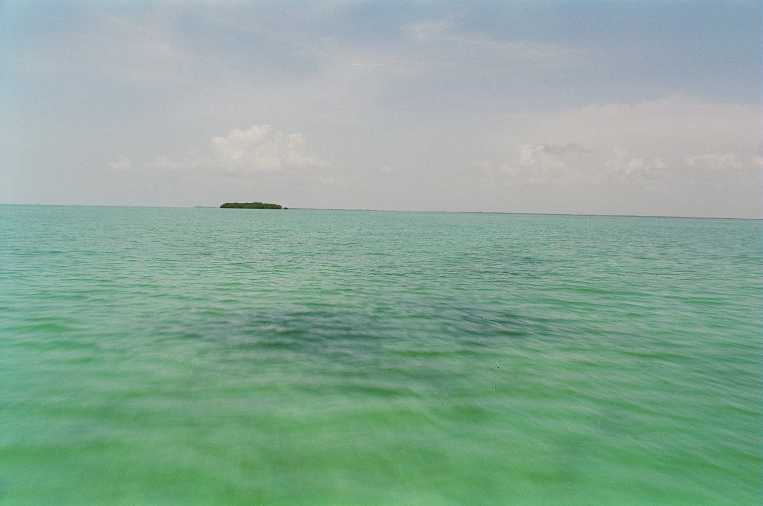 A small low island on the horizon surrounded by turquoise water under a hazy sky.