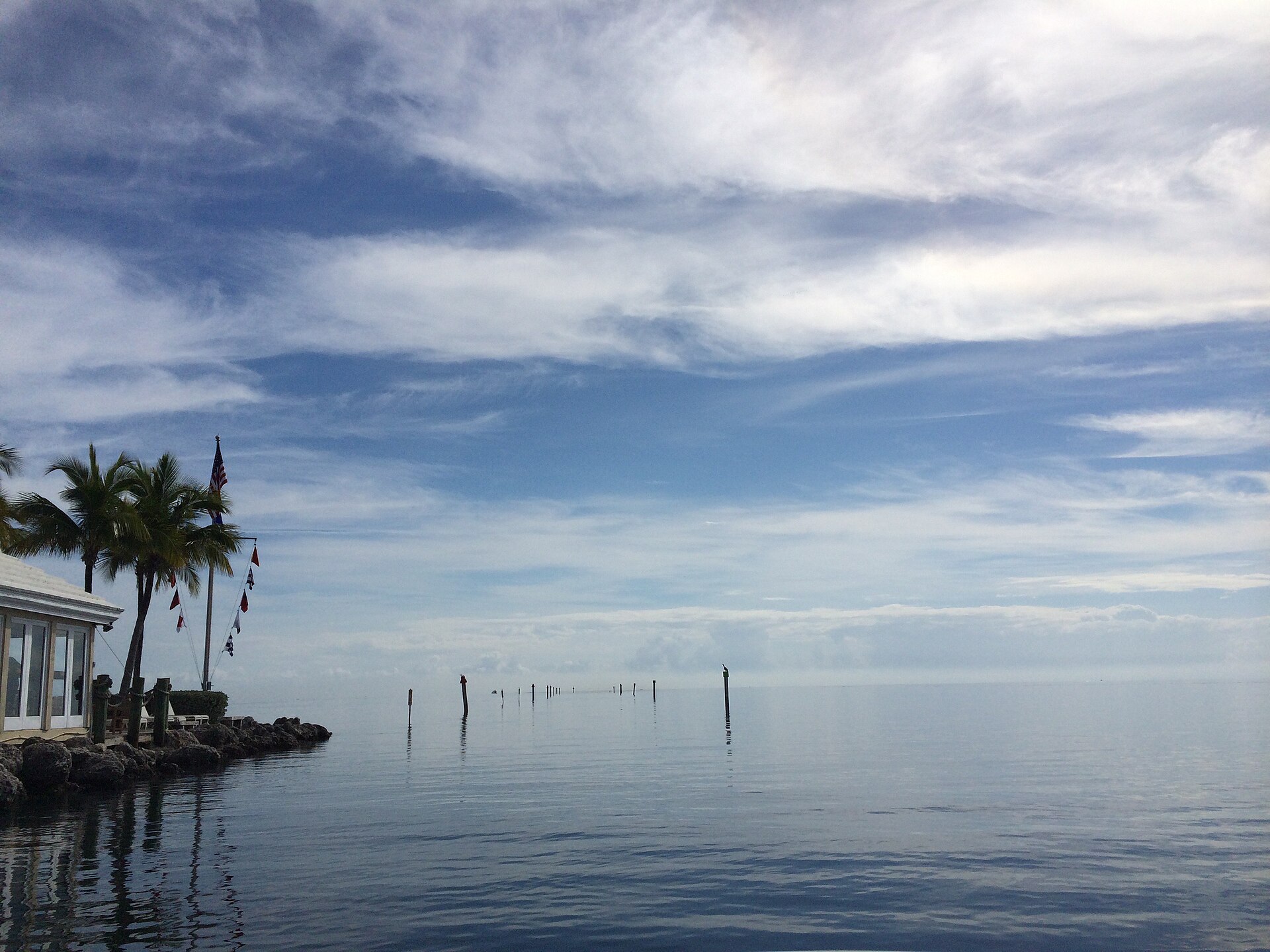 A waterfront club building with palm trees beside calm blue water dotted with channel markers under a bright sky.