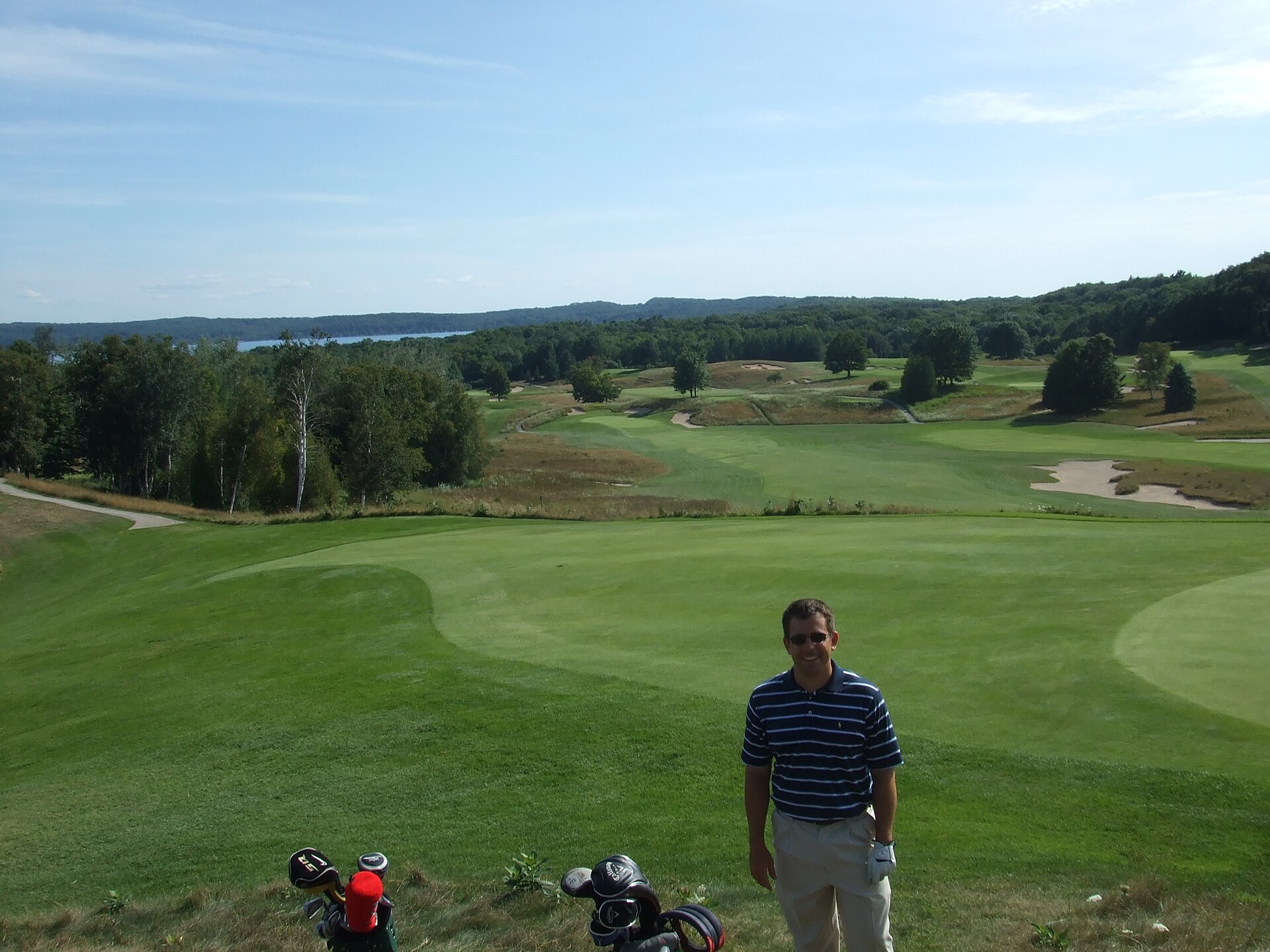 A golfer standing on a manicured green with rolling fairways, bunkers, and wooded bluffs behind.