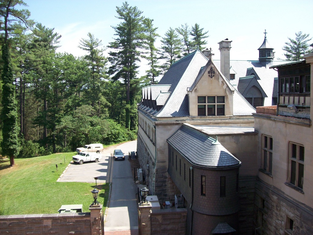 Stone chateau-style mansion wings with slate roofs, tall trees, and a service court with parked cars.