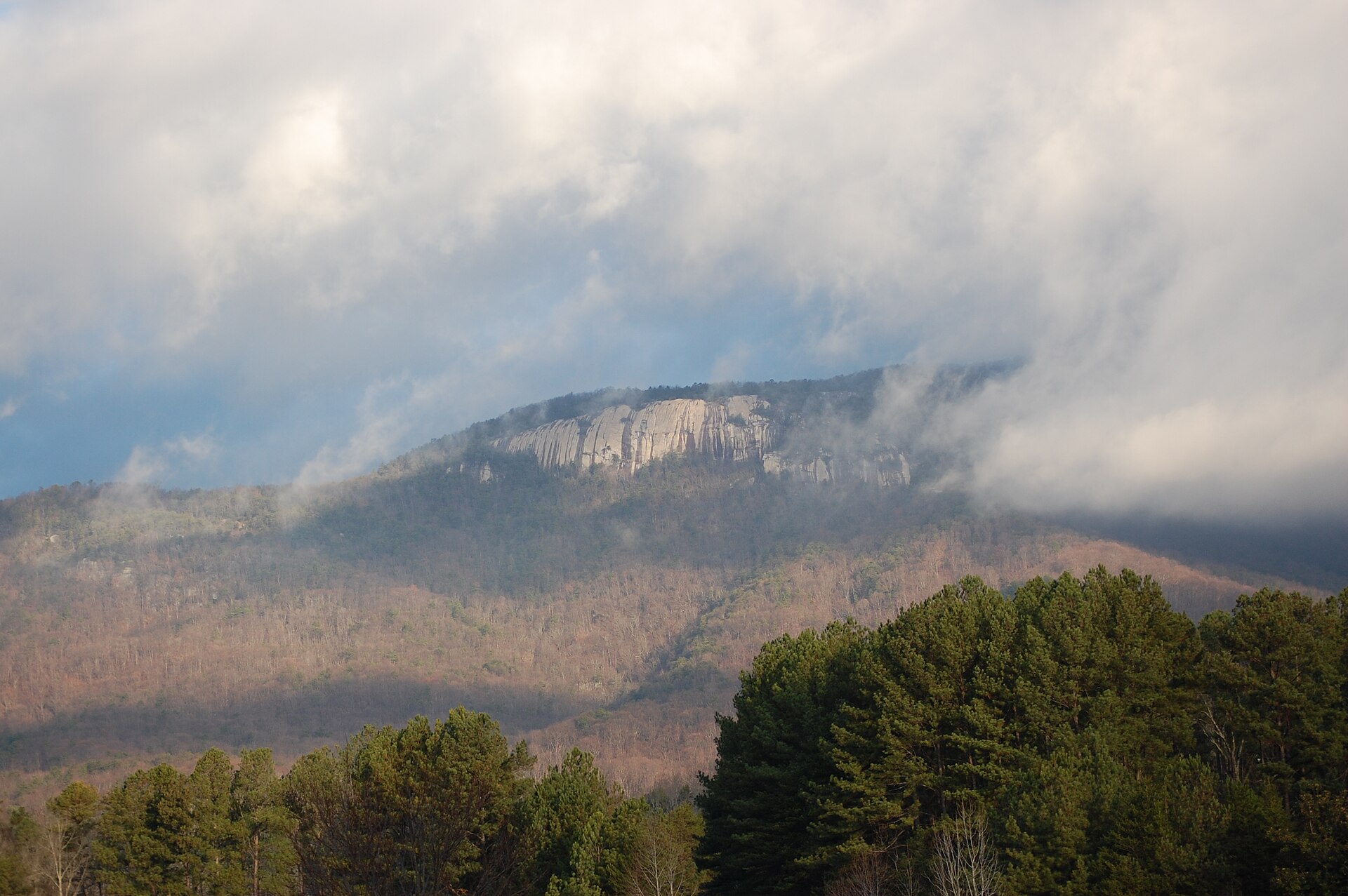 A sheer pale rock mountain face rising above forested slopes under partly cloudy skies.