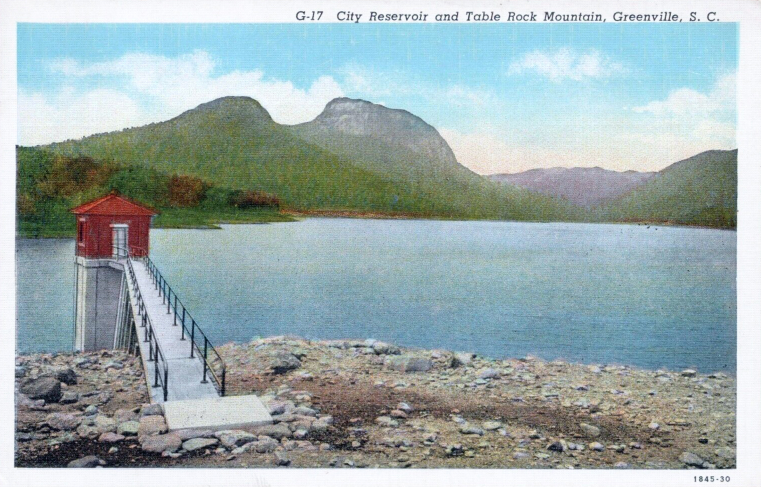 A hand-tinted vintage postcard showing a lake and small pumphouse with a mountain behind.