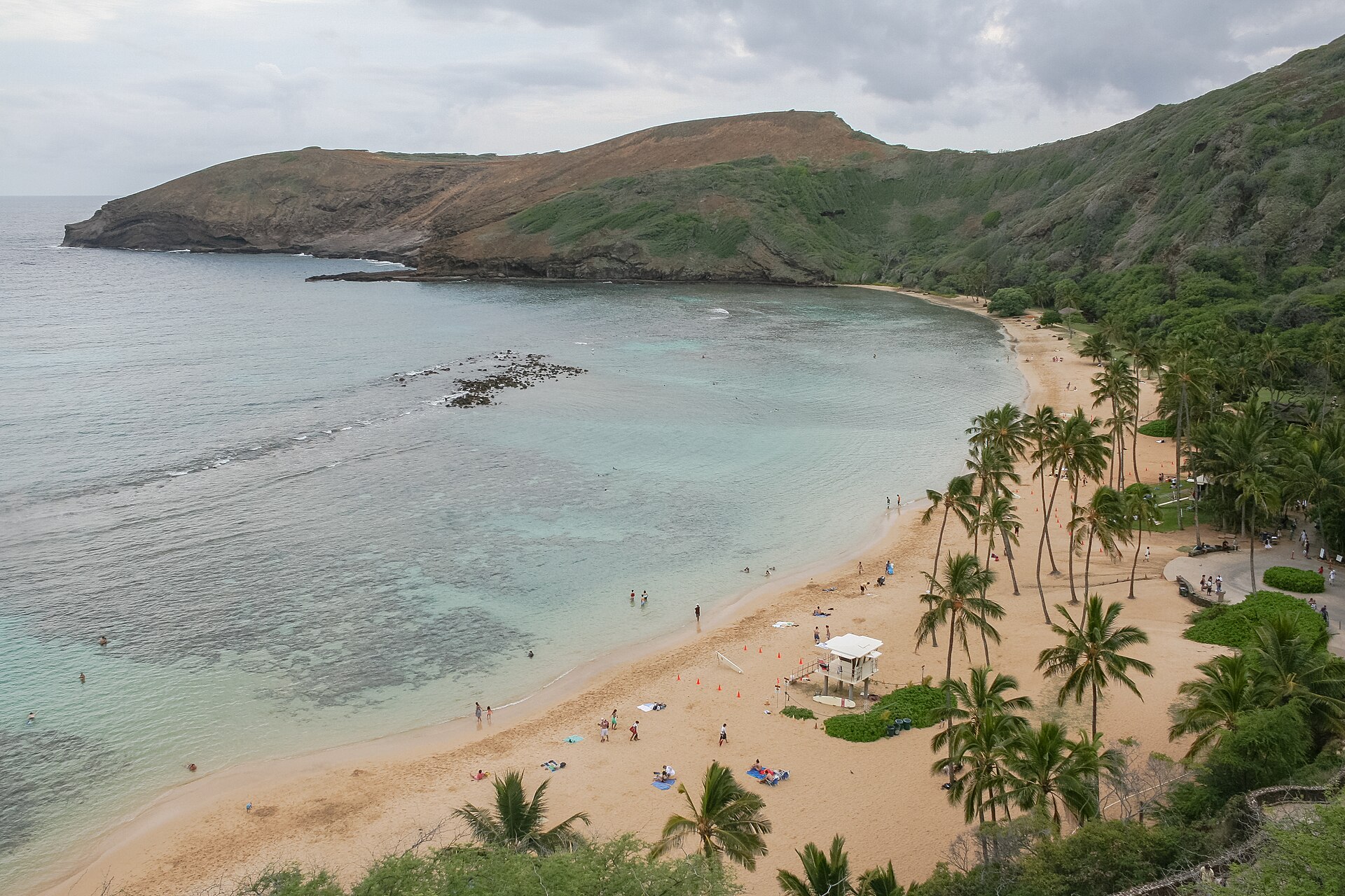 A curved sandy beach with palm trees and swimmers, ringed by a volcanic crater ridge and turquoise water.