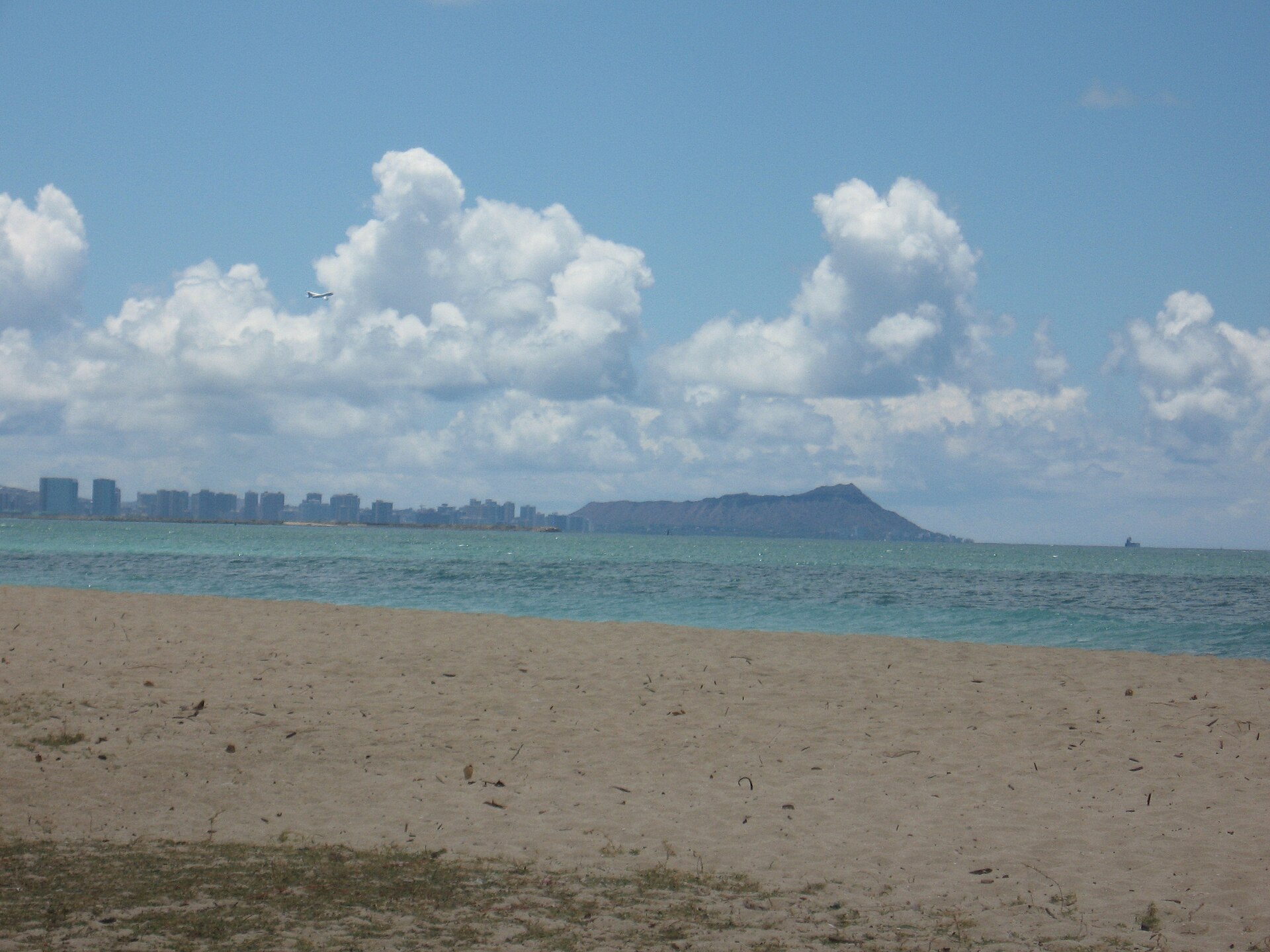 Sandy beach with palm trees and the distant silhouette of Diamond Head across blue water under a partly cloudy sky.