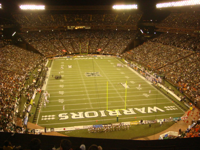 The interior of a large stadium with red and blue seats around a field, with advertising banners.
