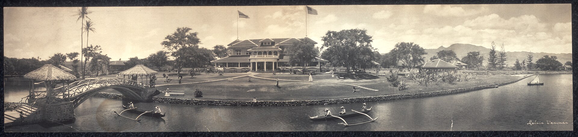 A sepia panoramic photograph of a grand beachfront hotel with palm trees and canoes on the sand.