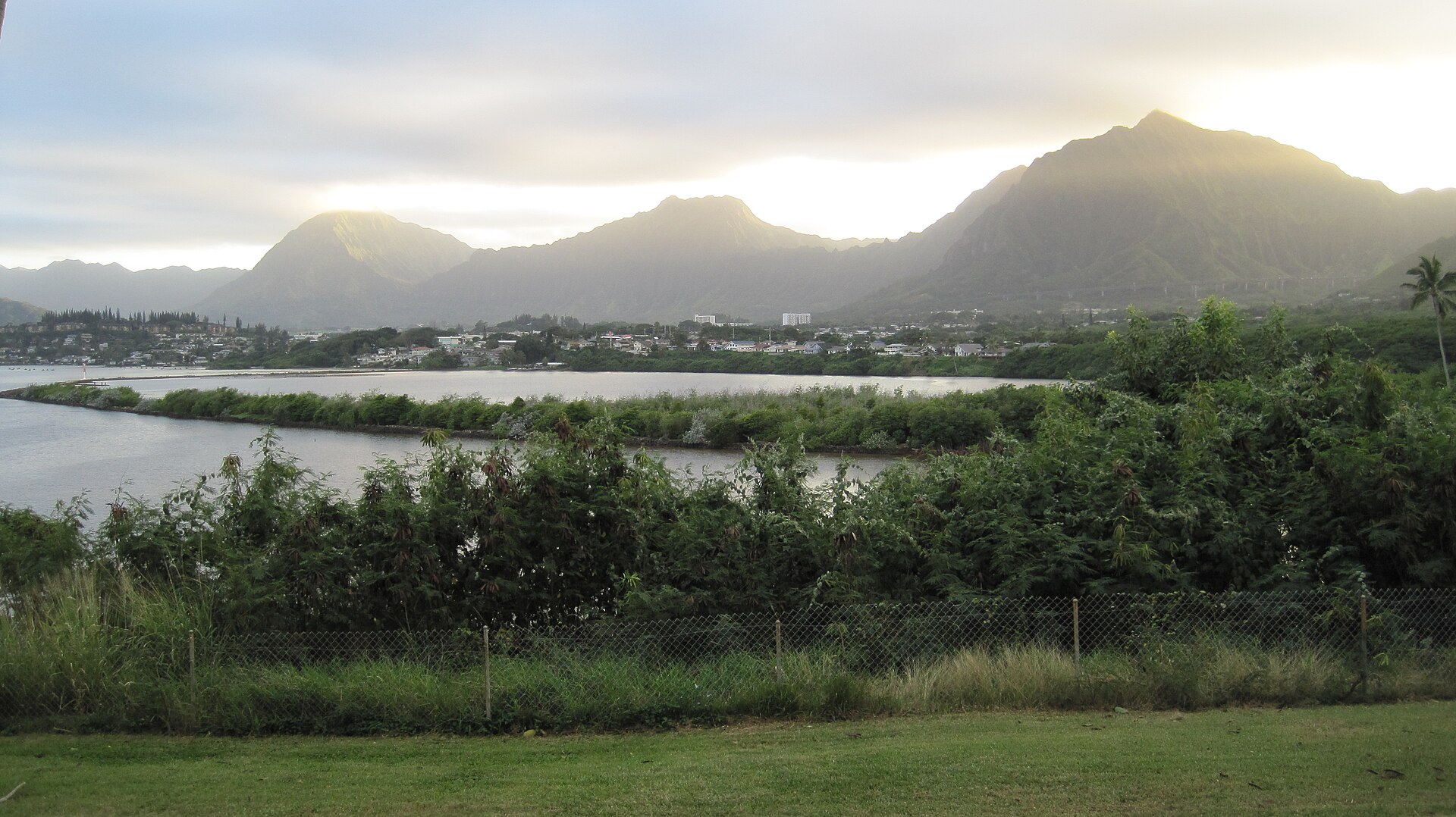 A long stone fishpond wall crossing calm water with steep green mountains rising behind.