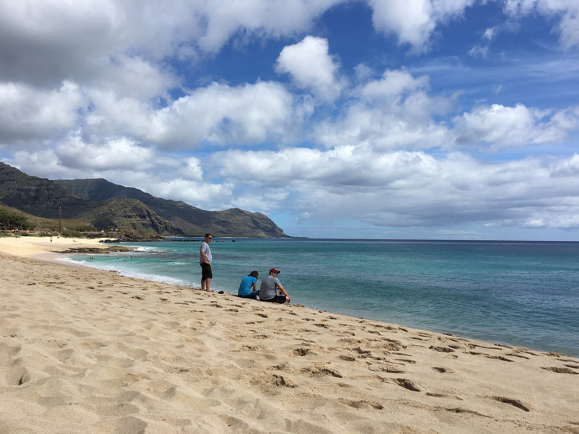 A wide sandy beach with turquoise water, beachgoers, and the Waianae mountains rising behind.