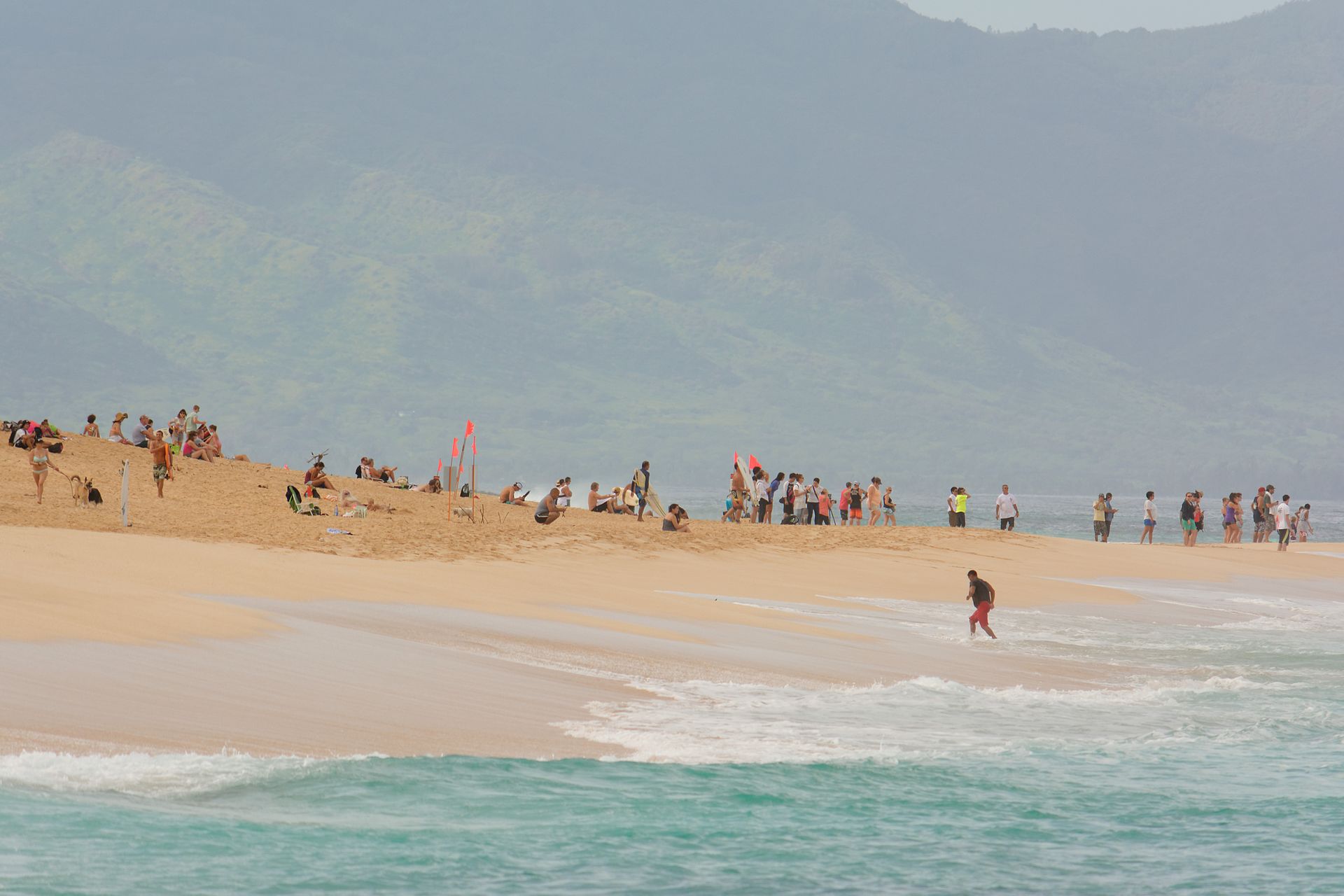 A crowded sandy beach with surfers and onlookers, turquoise waves, and hazy mountains behind.