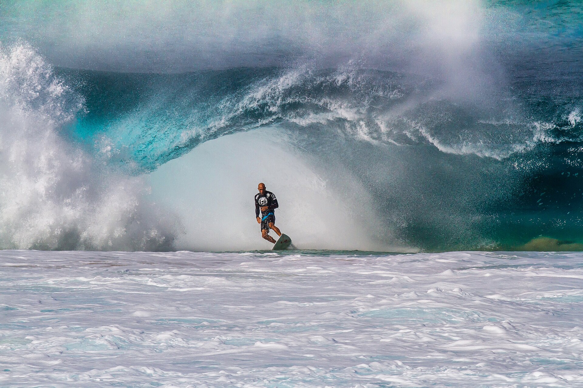 A surfer dwarfed inside a huge curling turquoise wave throwing spray.
