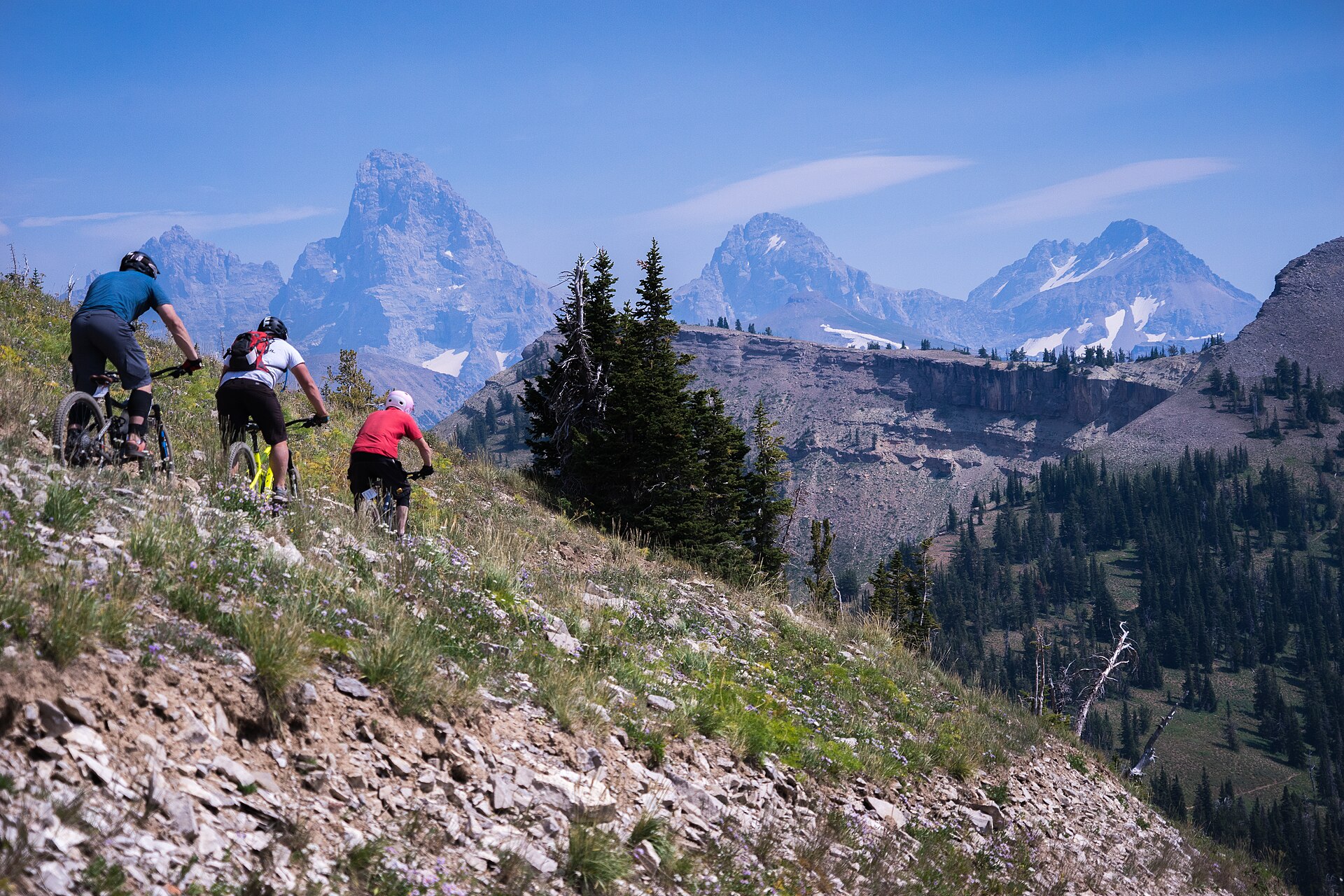Three mountain bikers descending a rocky trail with the Grand Teton and sister peaks towering behind.
