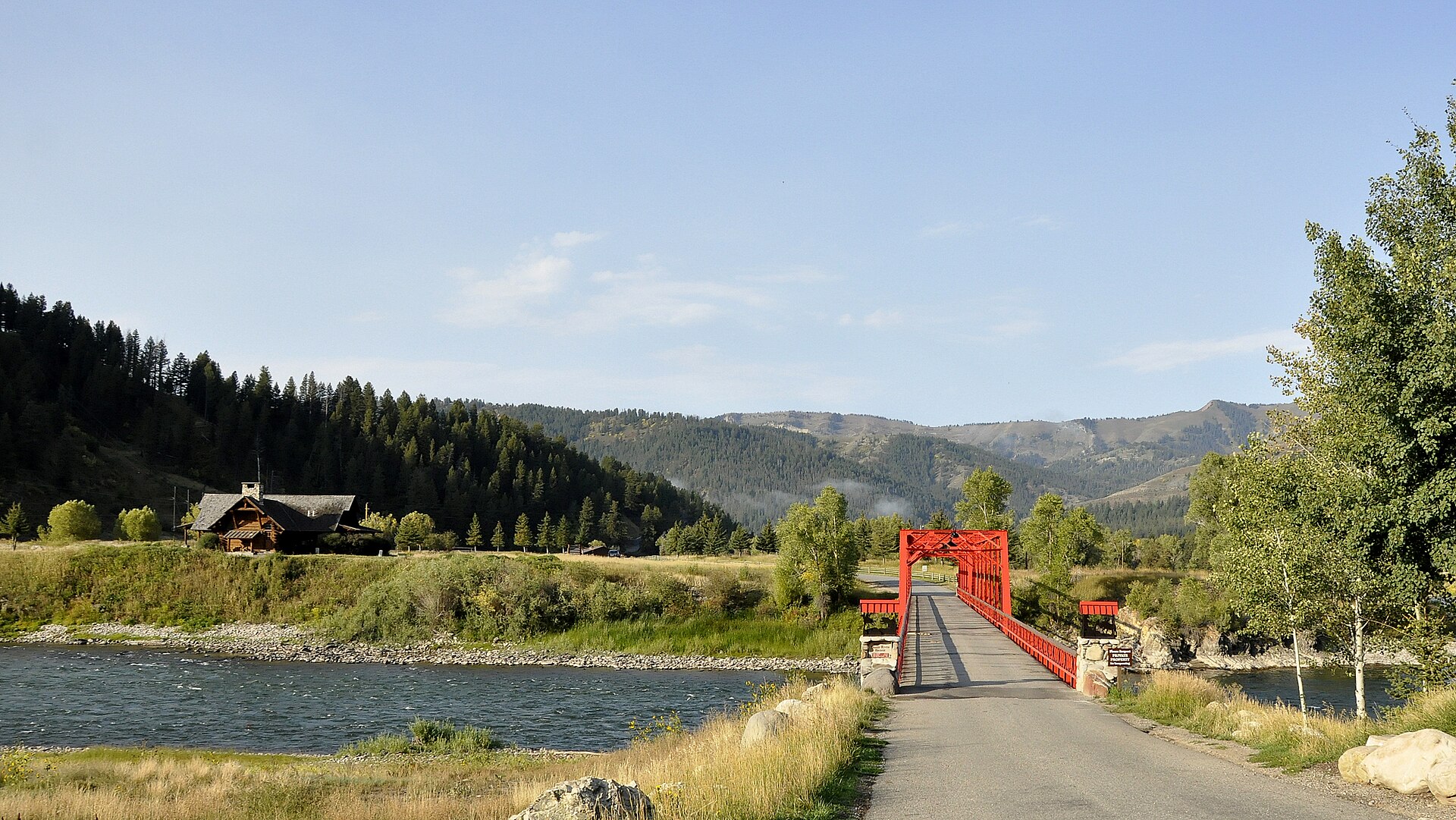 A red steel truss bridge crossing a wide river, with a log building, cottonwoods, and forested mountains.