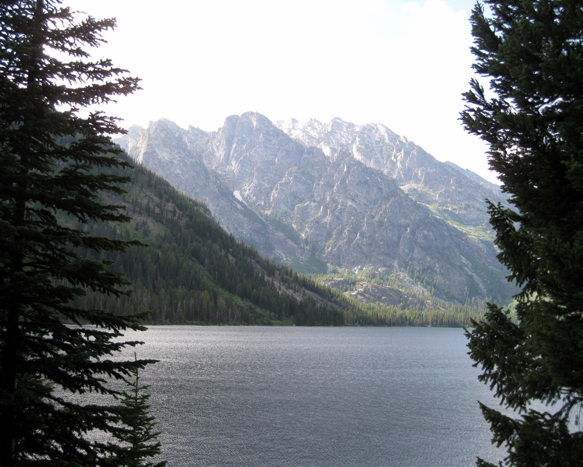 A calm lake framed by evergreens with rugged gray peaks rising behind.