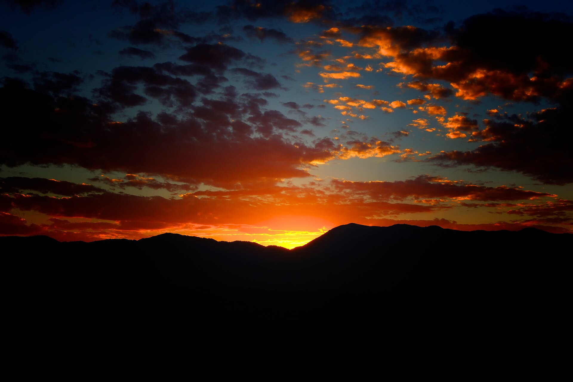 A fiery orange sunrise with dramatic clouds silhouetting dark mountain ridges.