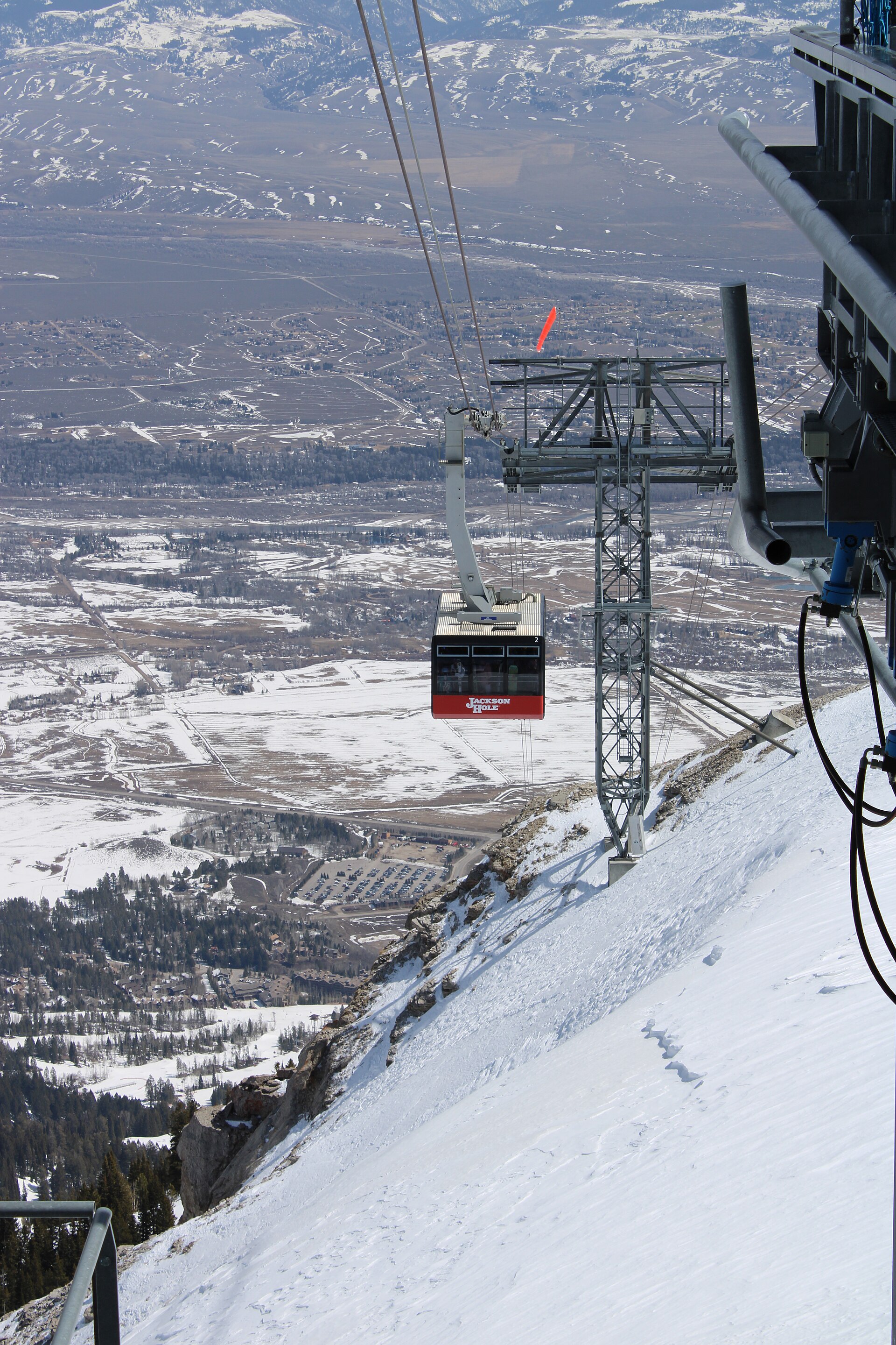 A red aerial tram car suspended from cables above a snowy mountainside, with the valley spread below.