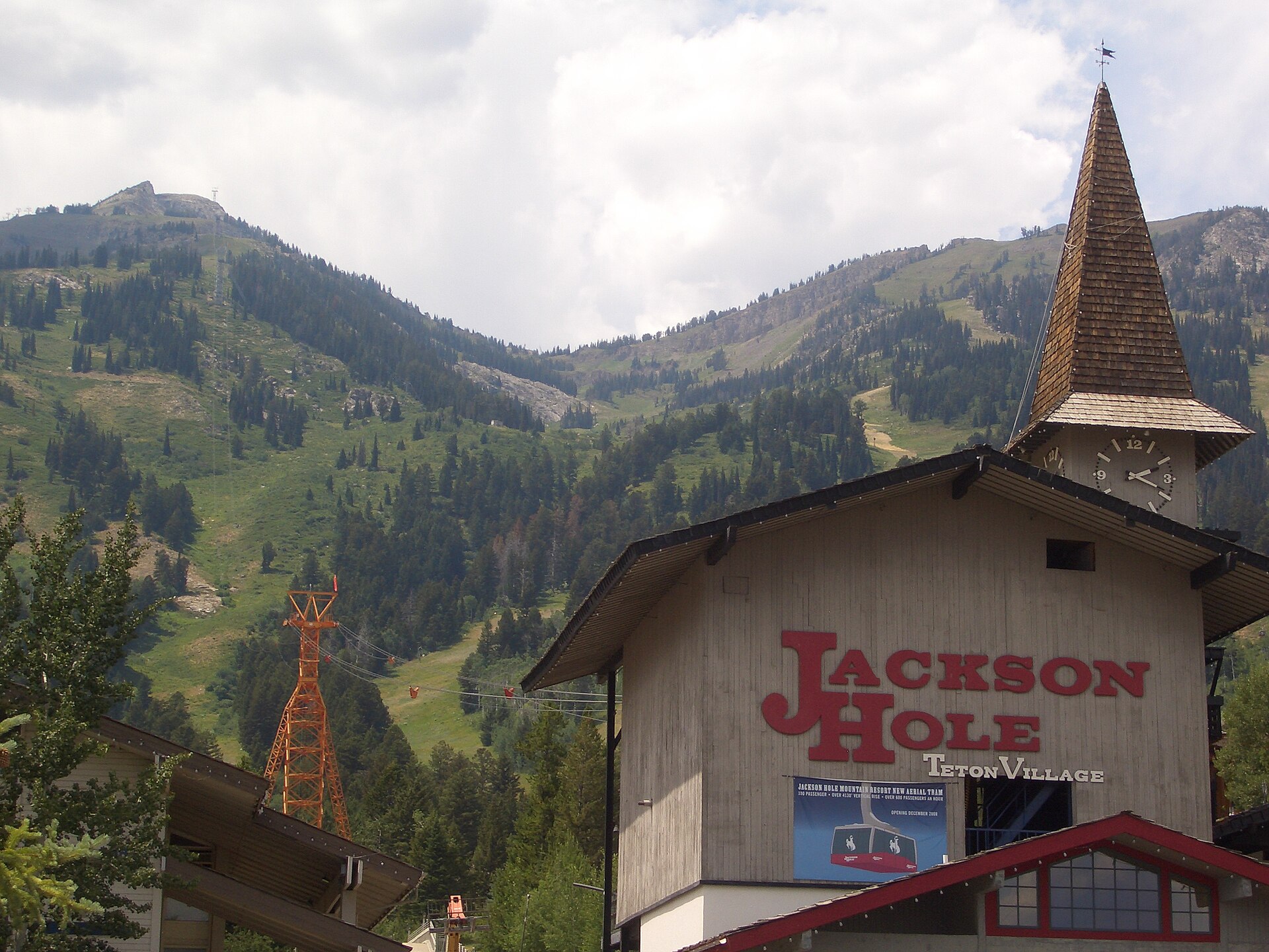 A wooden clock tower and station buildings with a 'Jackson Hole Teton Village' sign, backed by forested slopes.
