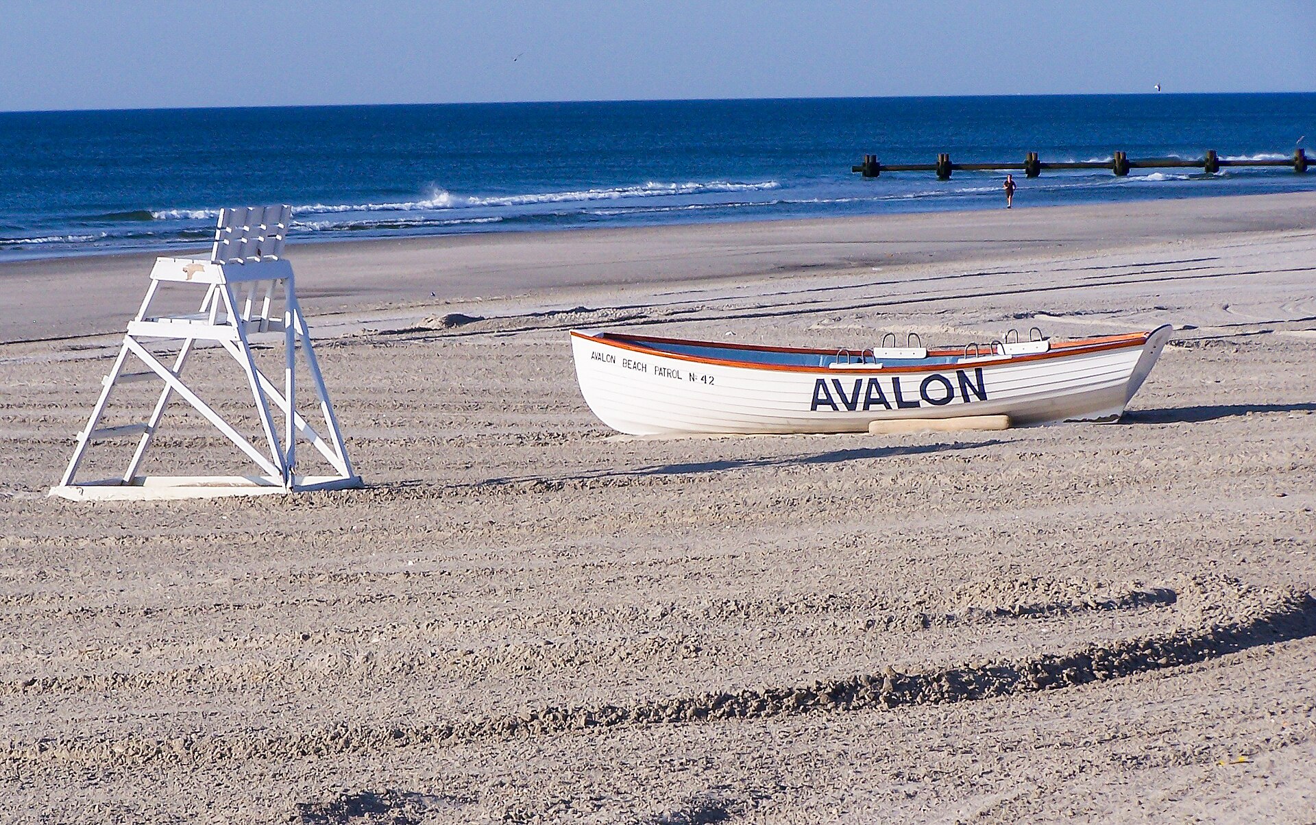 A white boat lettered 'AVALON' on a wide sandy beach beside a lifeguard stand, with the ocean beyond.