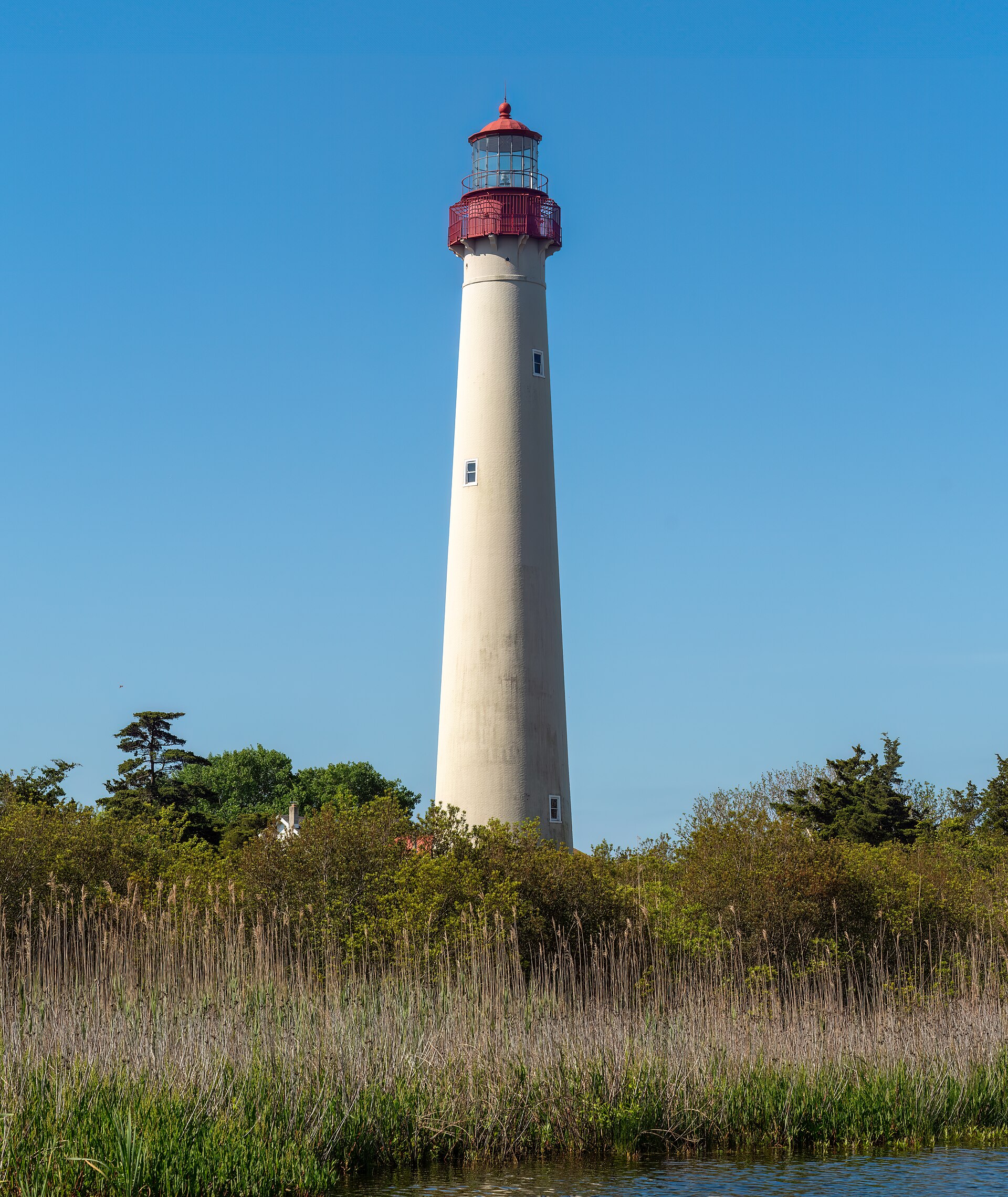 A tall white lighthouse with a red lantern room rising above marsh grasses under a blue sky.