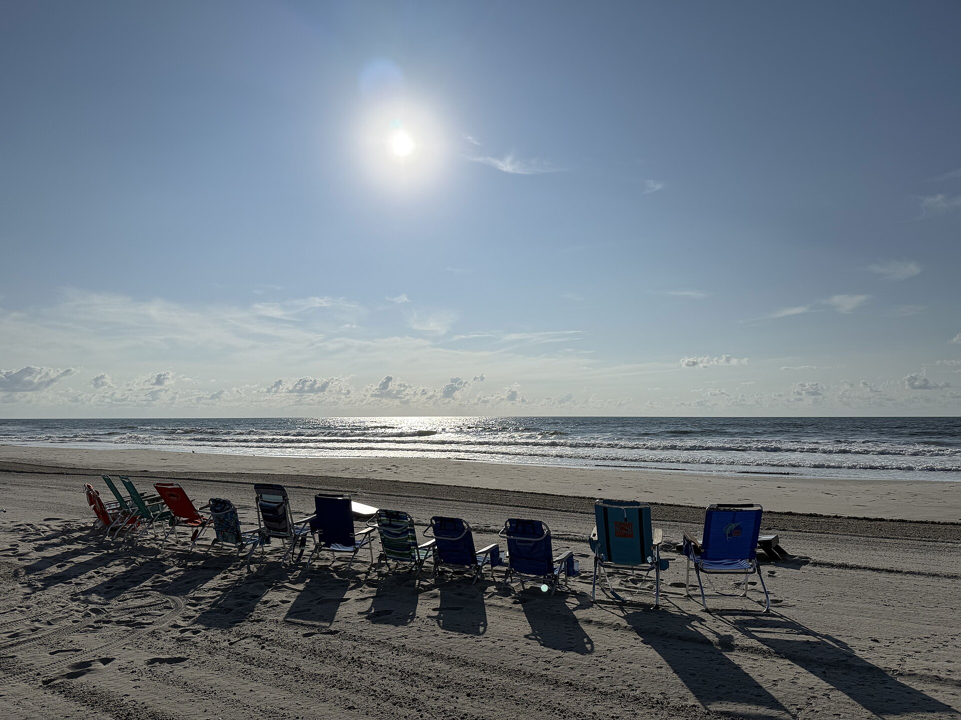 Empty beach chairs facing the ocean under a bright sun on a wide sandy beach.