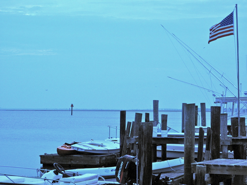 Fishing boats and wooden pilings at a quiet marina with an American flag, bathed in blue evening light.