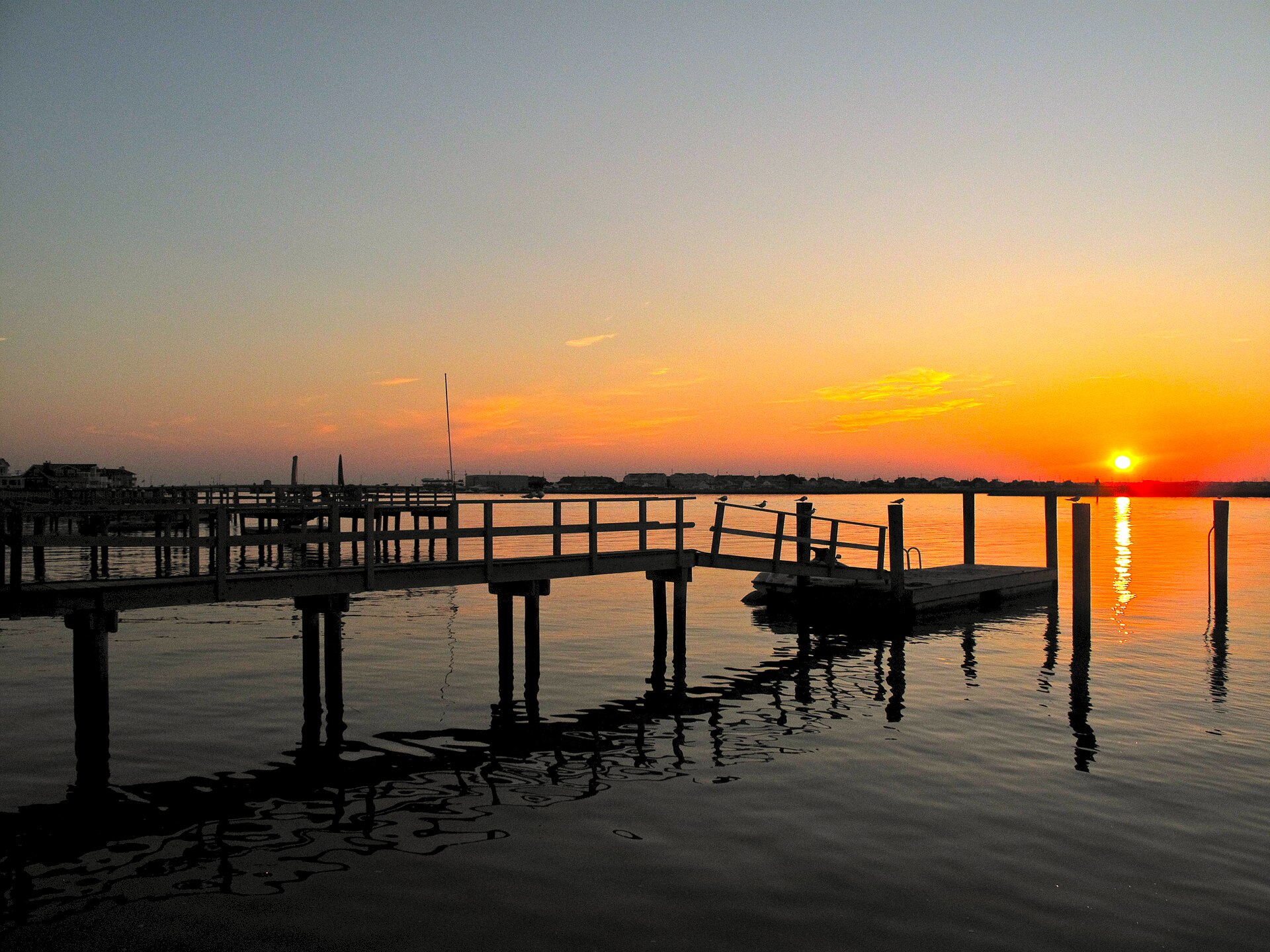 A wooden dock silhouetted against an orange sunset over calm bay water.