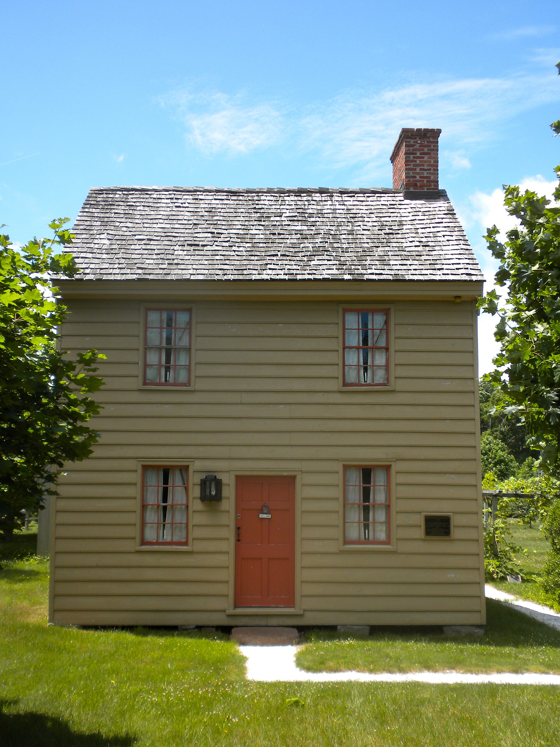 A small historic two-story clapboard house with a red door and brick chimney on a green lawn.