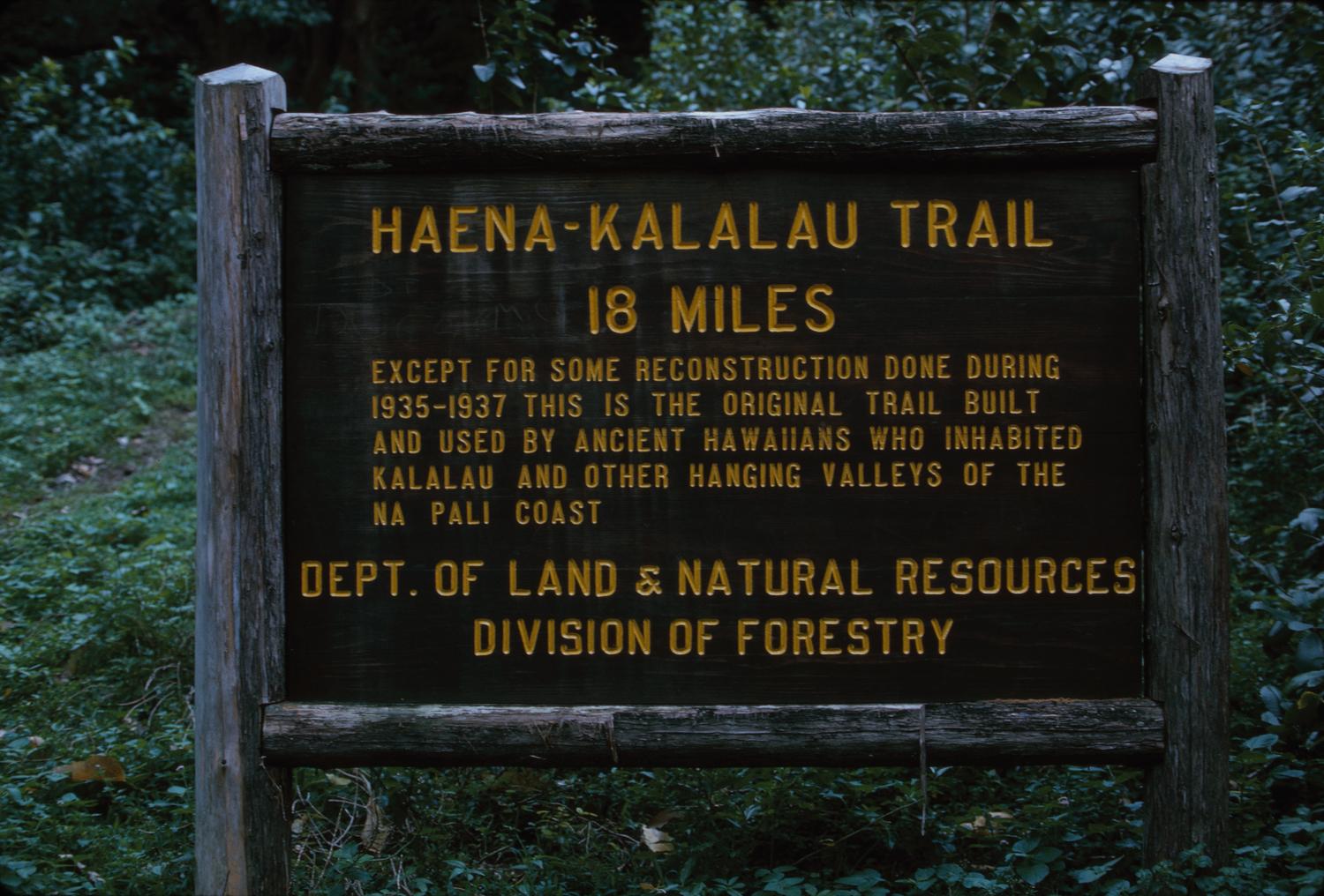 A brown wooden sign reading 'HAENA-KALALAU TRAIL 18 MILES' surrounded by green foliage.