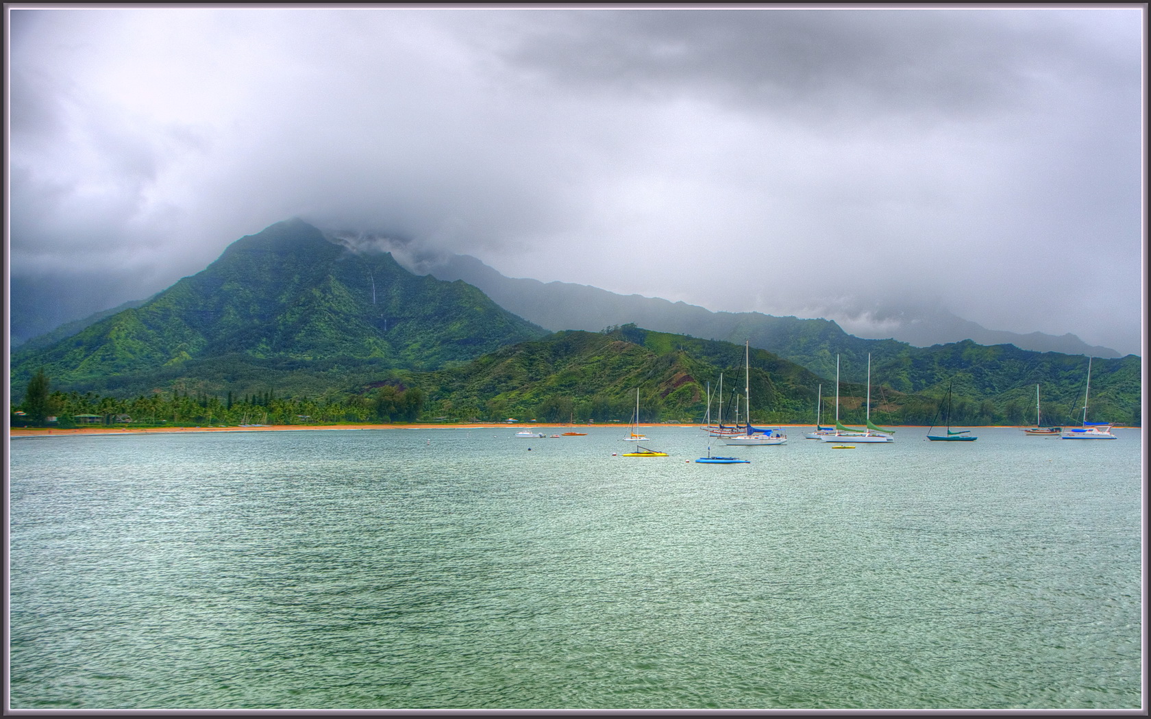 Sailboats on calm greenish bay water with steep cloud-draped mountains rising behind.