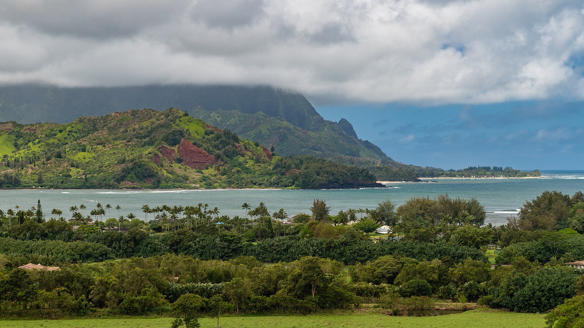 A wide bay with gentle surf, backed by lush green ridges under a cloudy sky.