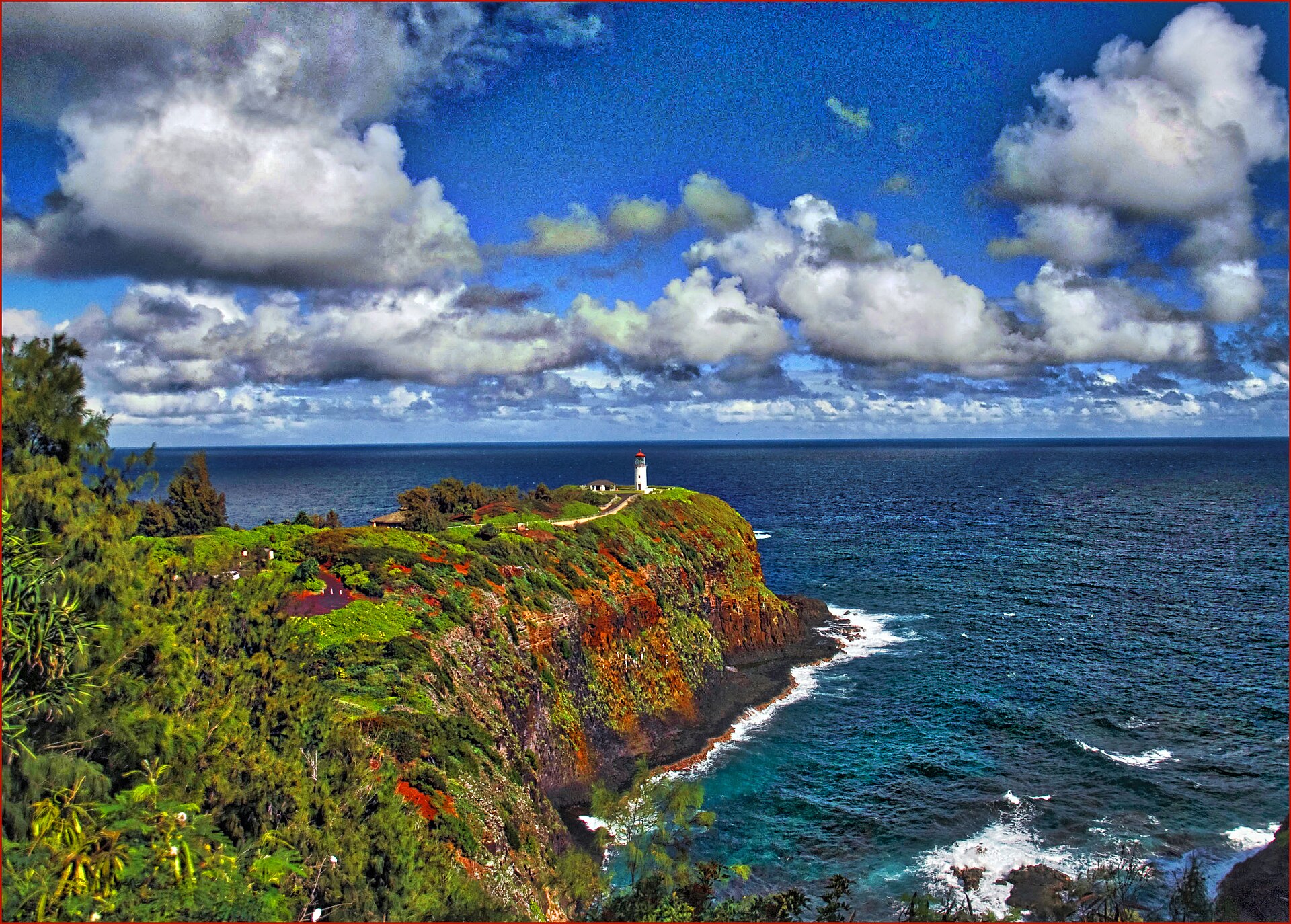 A white lighthouse with a red roof on a lush green point above deep blue ocean, under dramatic clouds.