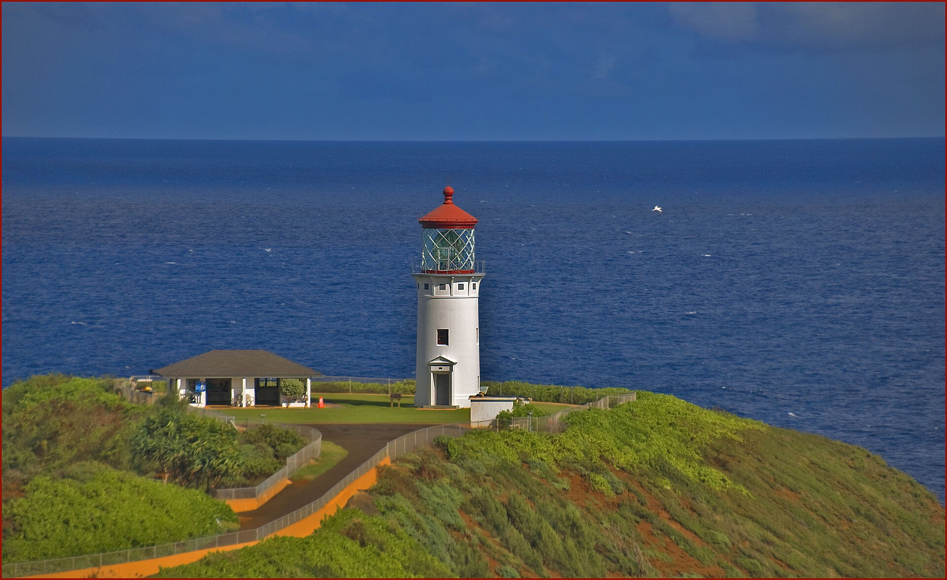 A white lighthouse with a red lantern room on a grassy bluff, with deep blue ocean stretching to the horizon.