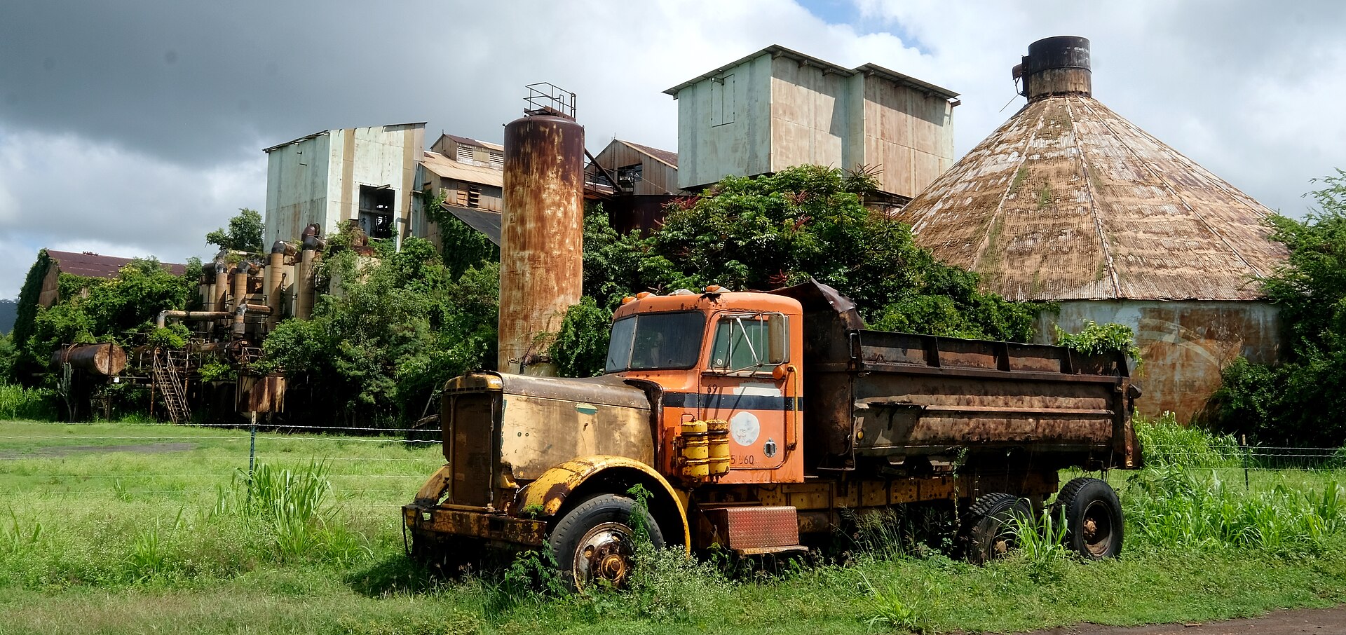 A rusting orange truck parked before the derelict concrete sugar mill buildings, overgrown with grass.
