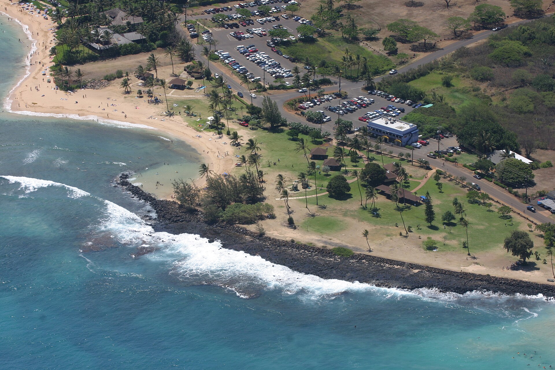 A curving sandy beach with a full parking lot, park buildings, and turquoise surf breaking on dark rock.