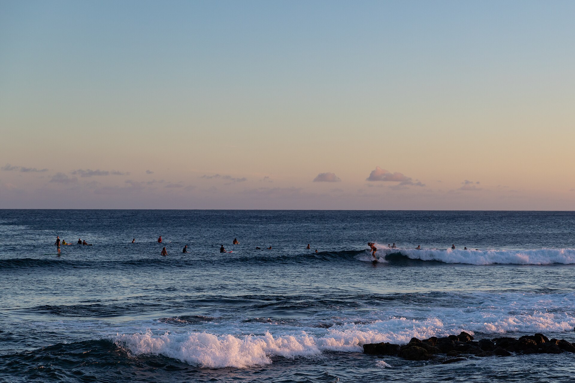 Surfers on gentle waves under a pastel sunset sky, with dark shoreline rocks in the foreground.