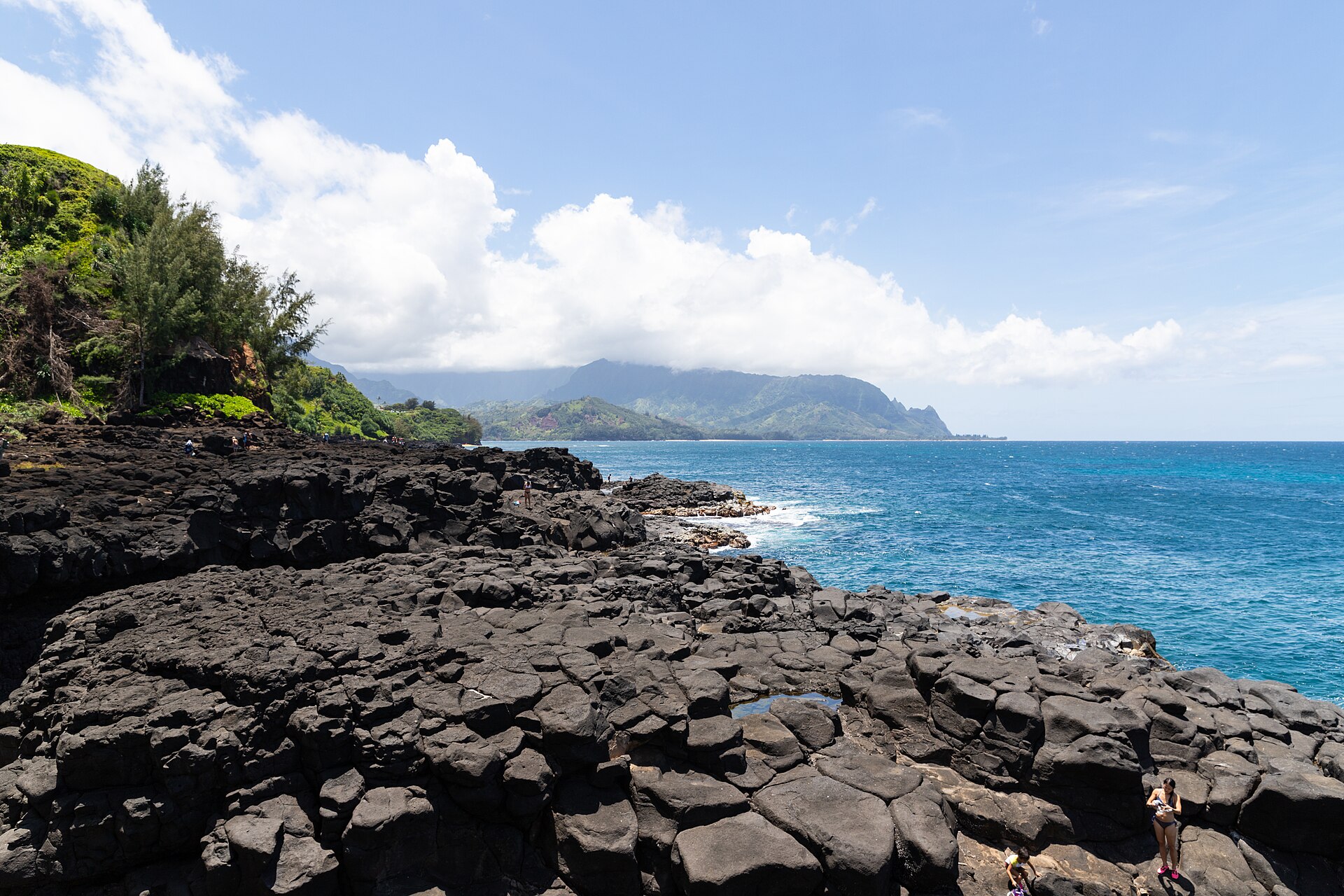 Black volcanic rock formations along a turquoise shoreline, with green mountains in the distance.