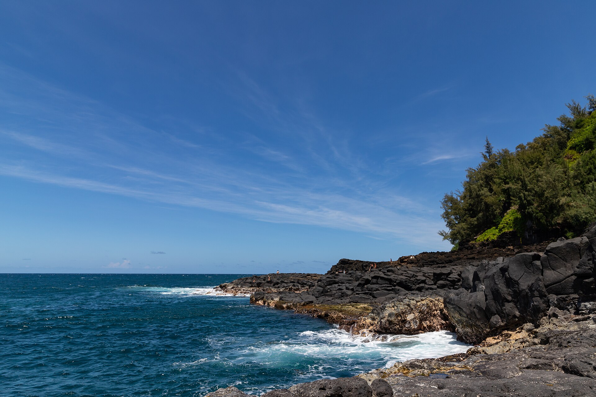 White surf breaking over dark lava rocks under a bright blue sky.