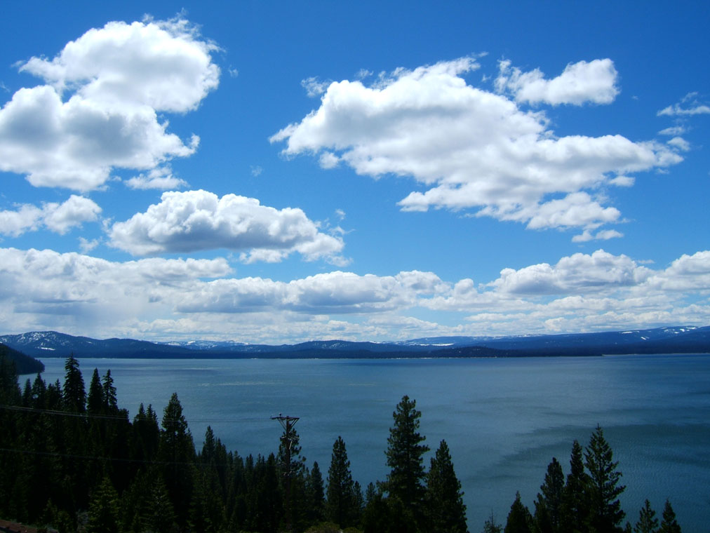 A blue lake with forested shoreline and mountains under a partly cloudy sky.