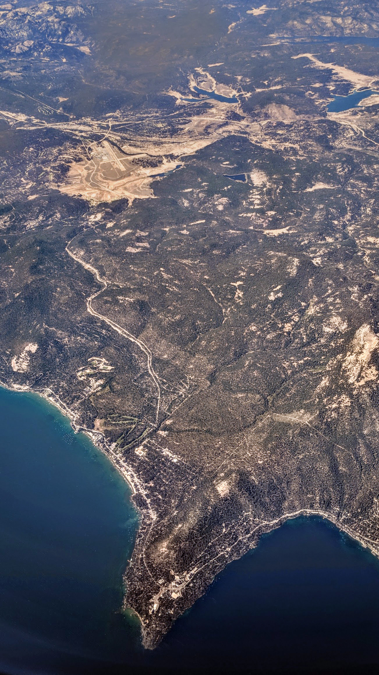 Aerial view of a forested peninsula with a curving shoreline, roads, and deep blue lake water on both sides.